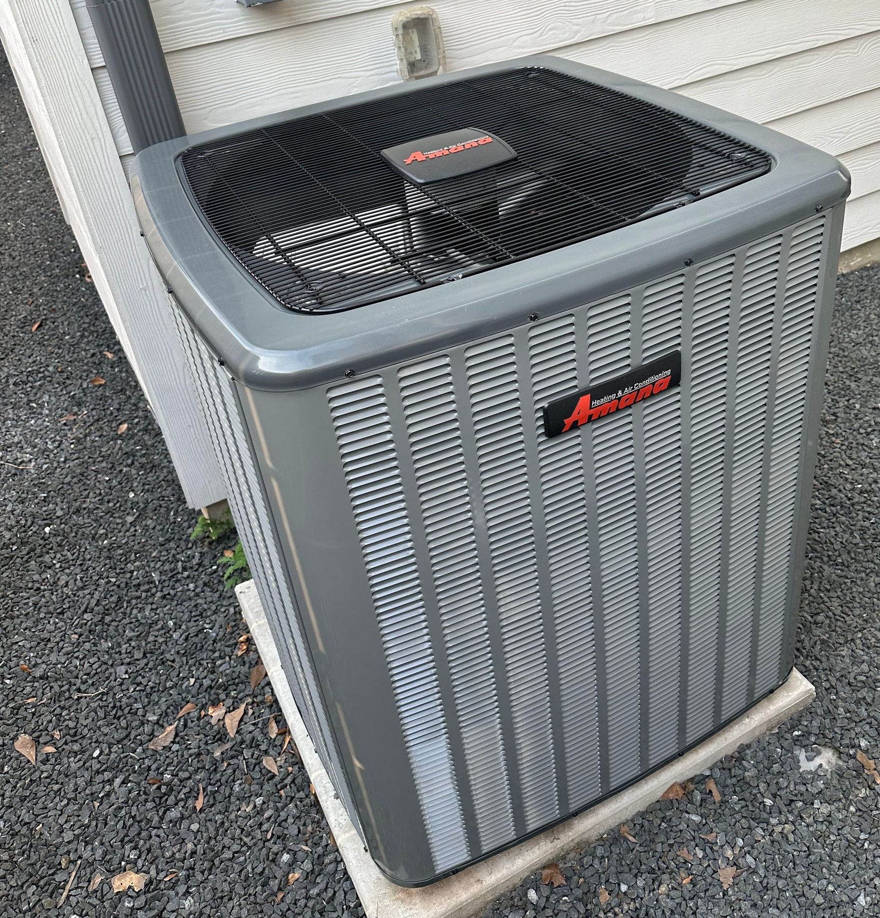 Gray Amana air conditioner unit outside against a white-sided house on a gravel base.