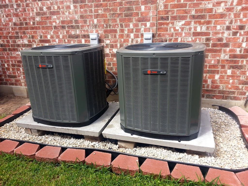 Two gray air conditioning units on concrete pads against a brick wall, bordered by red bricks and white gravel.