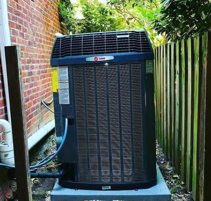 A dark HVAC unit between a brick wall and a wooden fence; situated on a gray platform.