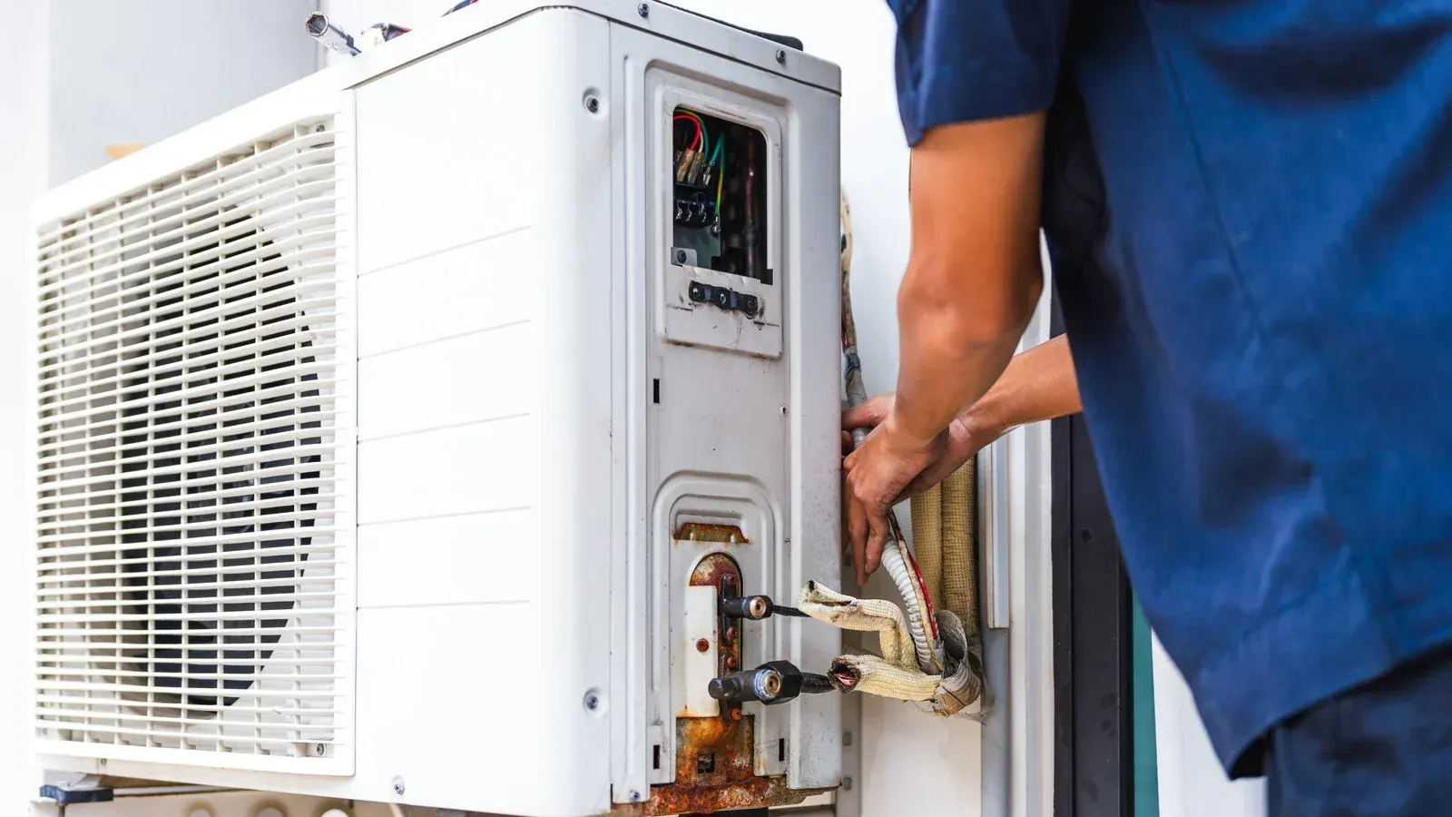 Person in blue shirt repairs outdoor AC unit, white and rusty.