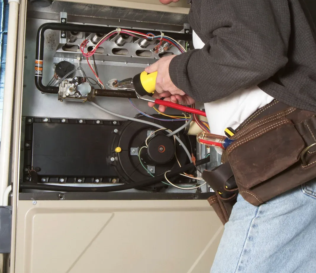 HVAC technician using a flashlight and screwdriver inside a furnace, likely for repair.
