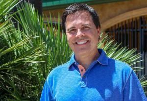 A man in a blue shirt is smiling in front of a palm tree.