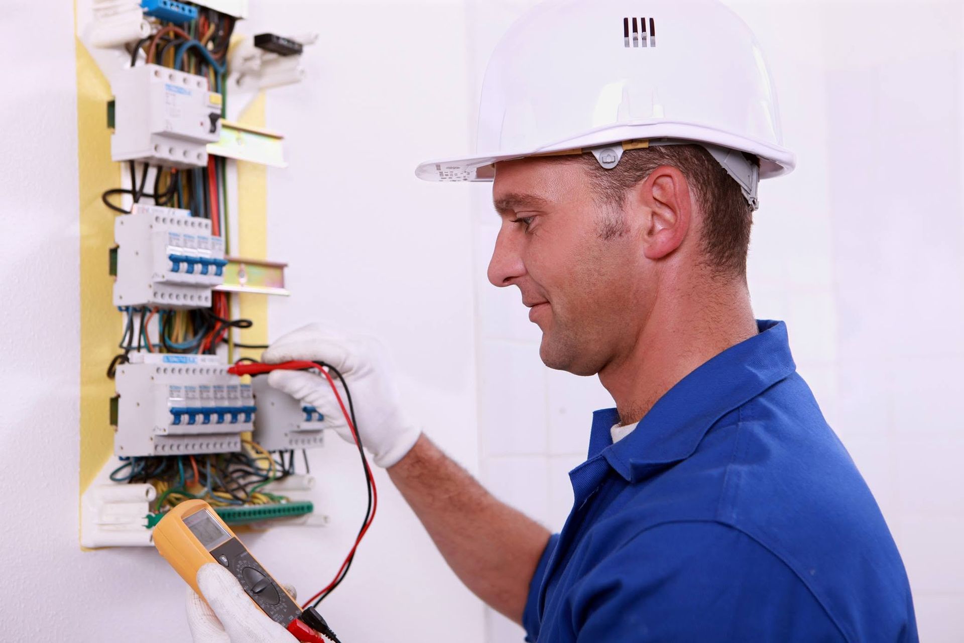 Electrician in white hard hat inspecting an open electrical panel with multimeter leads attached