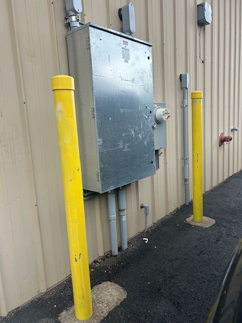 Yellow bollards beside a wall-mounted metal electrical box on a beige industrial building.