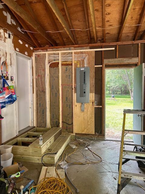 Unfinished garage wall with electrical panel, stacked lumber, ladder, and cords on concrete floor.