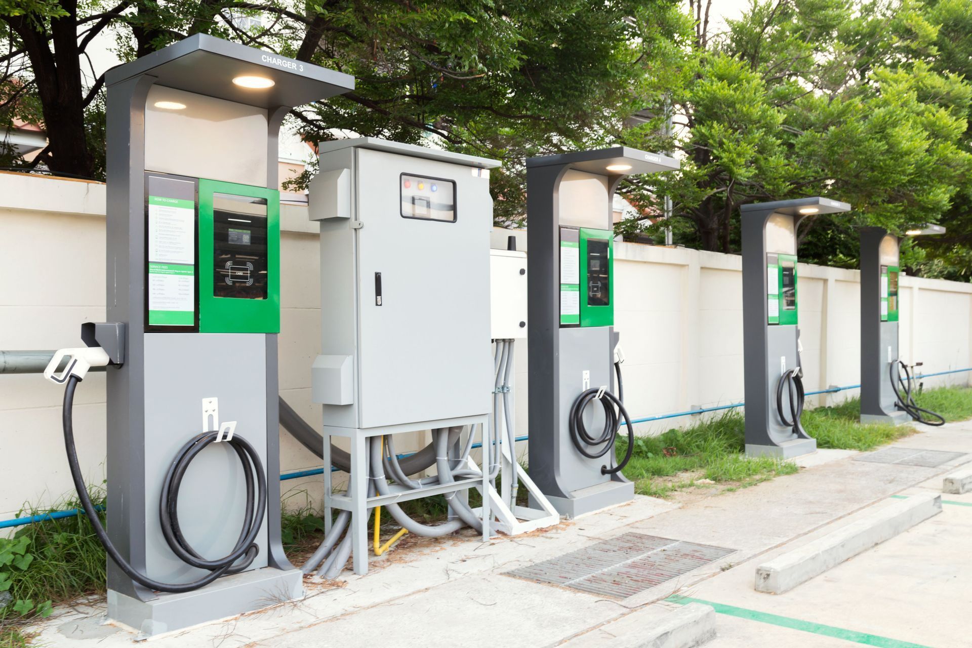 Row of electric vehicle charging stations with green-lit panels along a sidewalk under trees