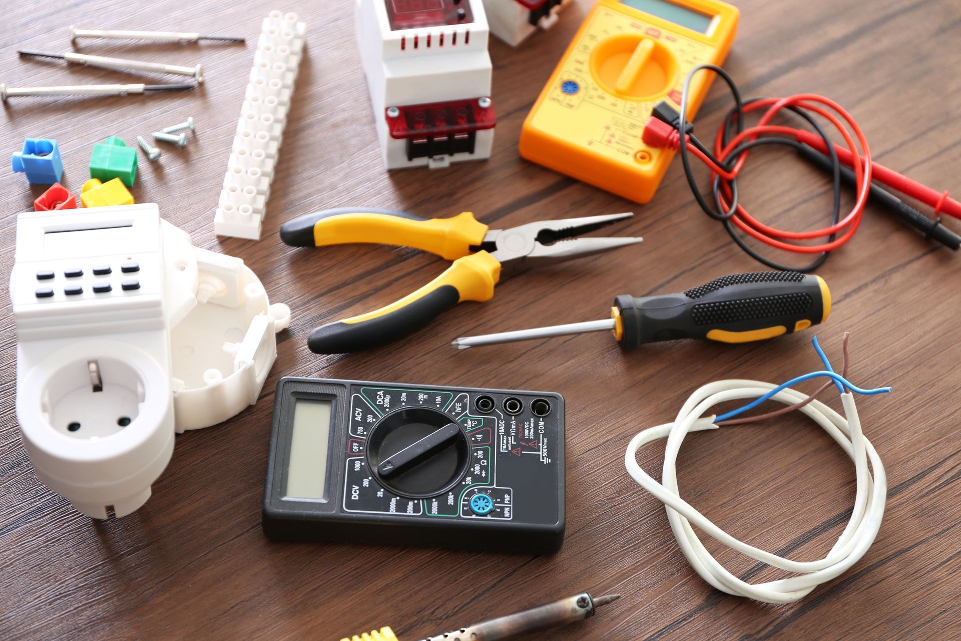 Electrical tools and outlets on a wooden surface, including pliers, a multimeter, wire, and adapters.