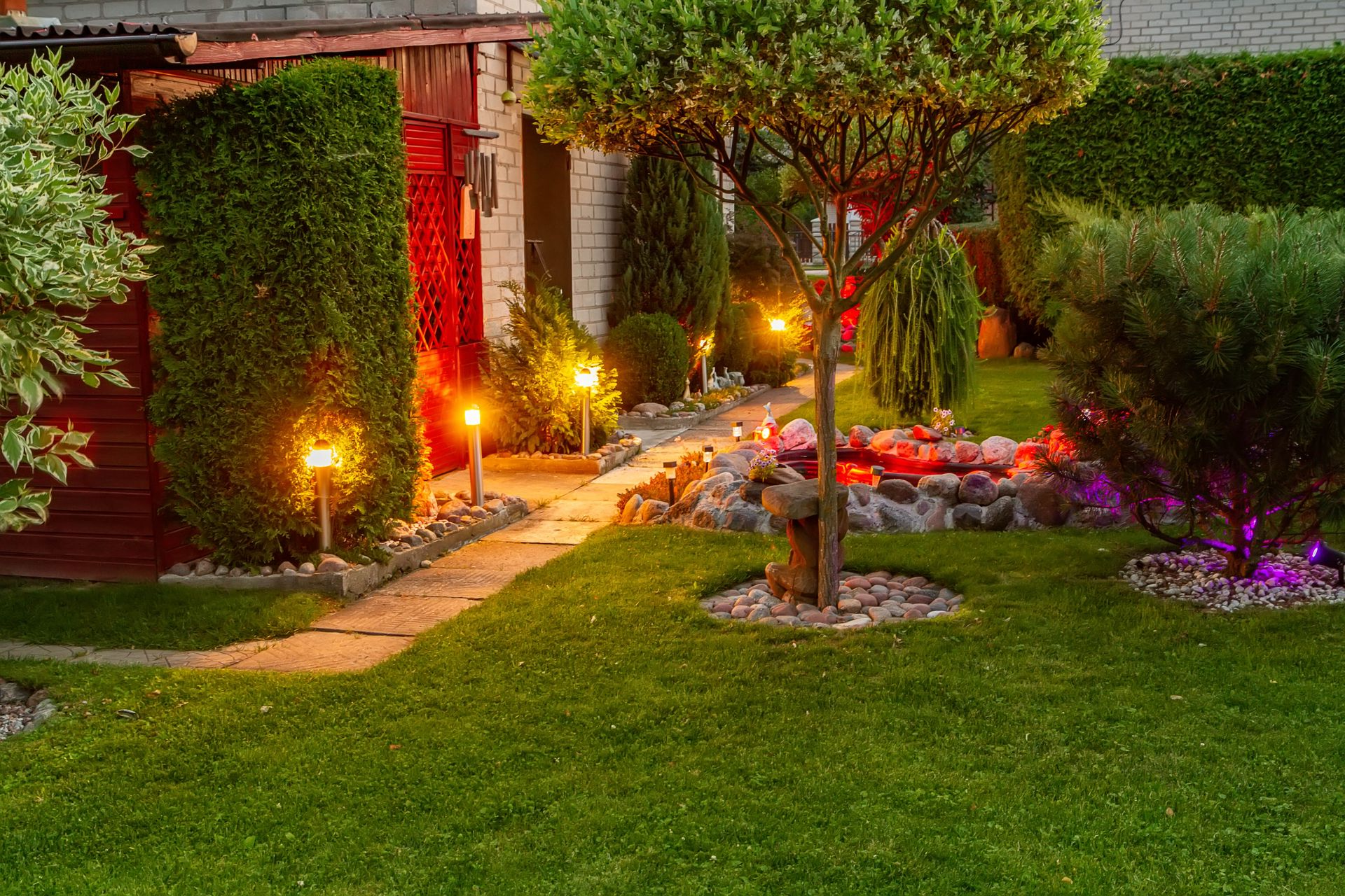Lit garden walkway beside a house, with warm path lights, flowers, trees, and a manicured green lawn.