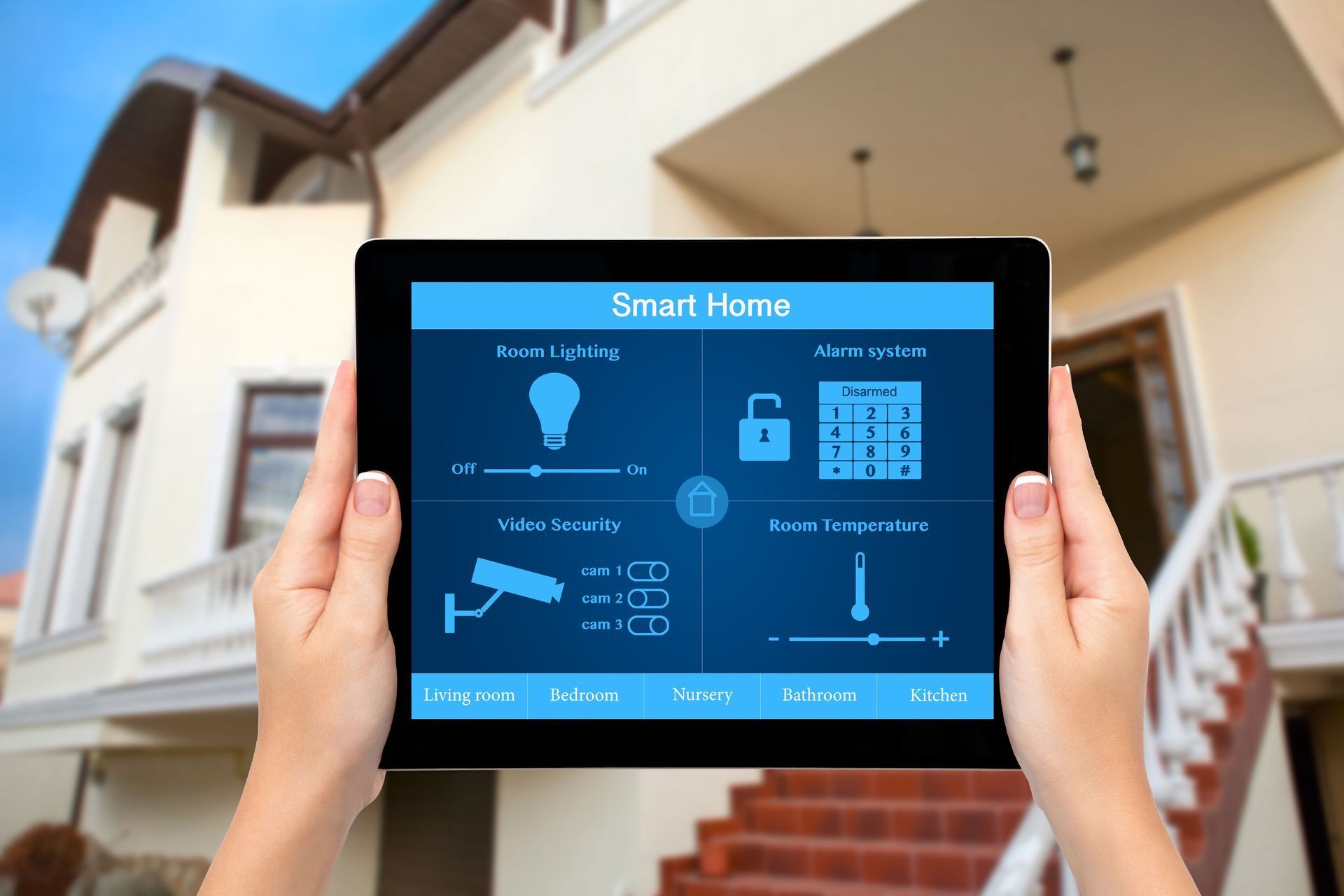Hands holding a tablet showing a smart home control dashboard in front of a house