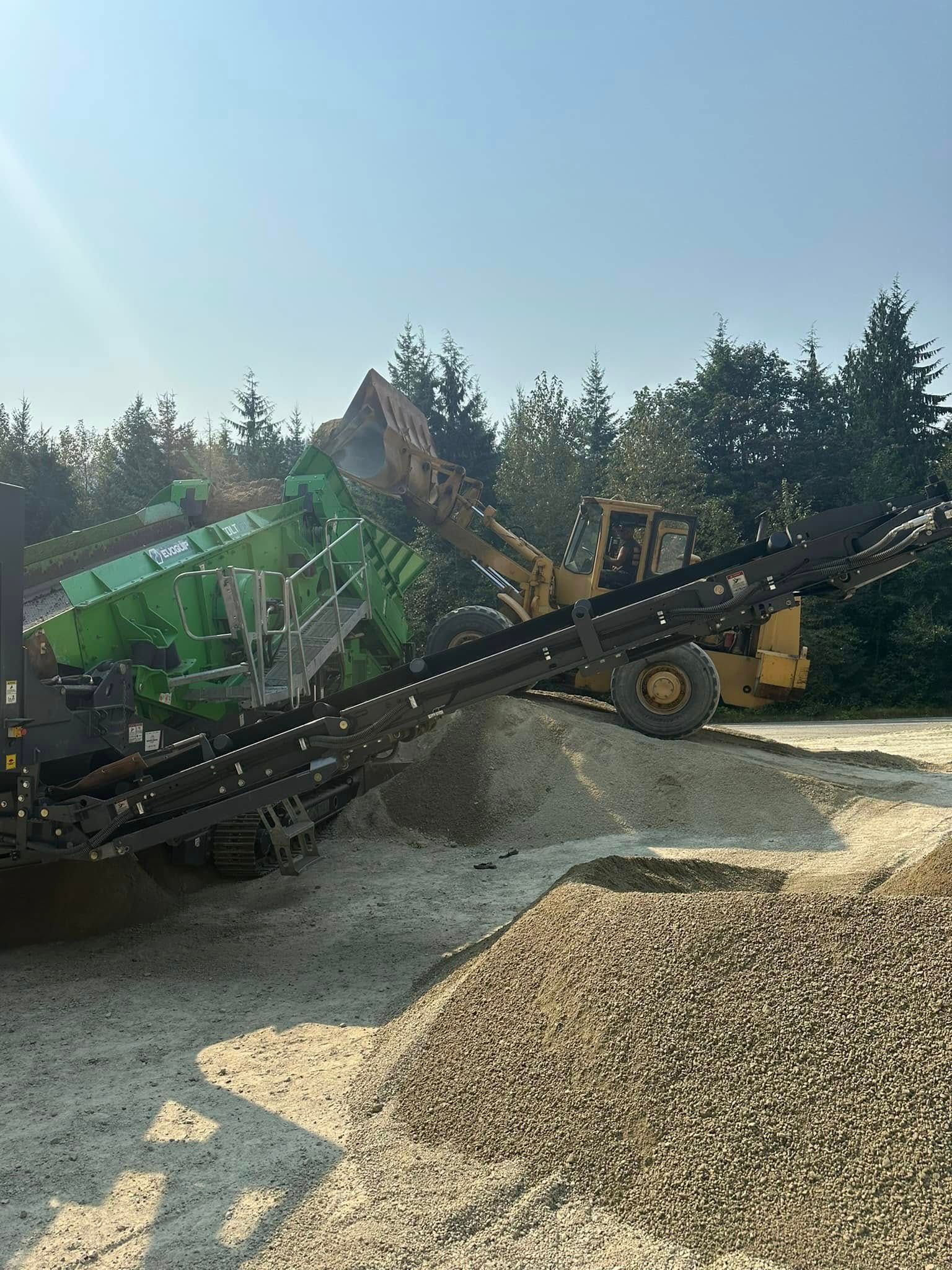 Yellow loader dumping material onto a conveyor belt; green machine in the background. Gravel and trees in the setting.