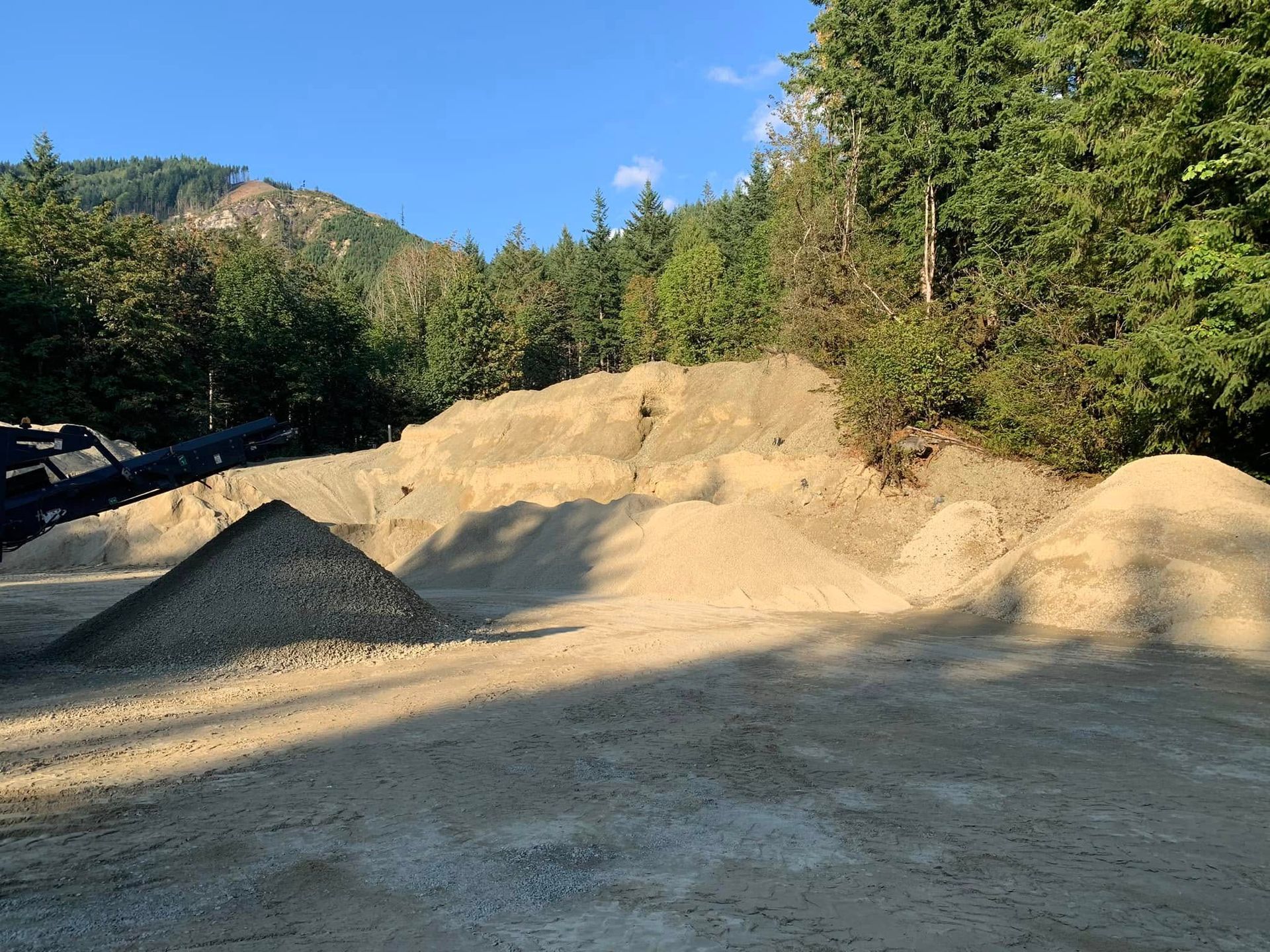 Piles of gravel and sand in a wooded area, with trees and a clear blue sky.