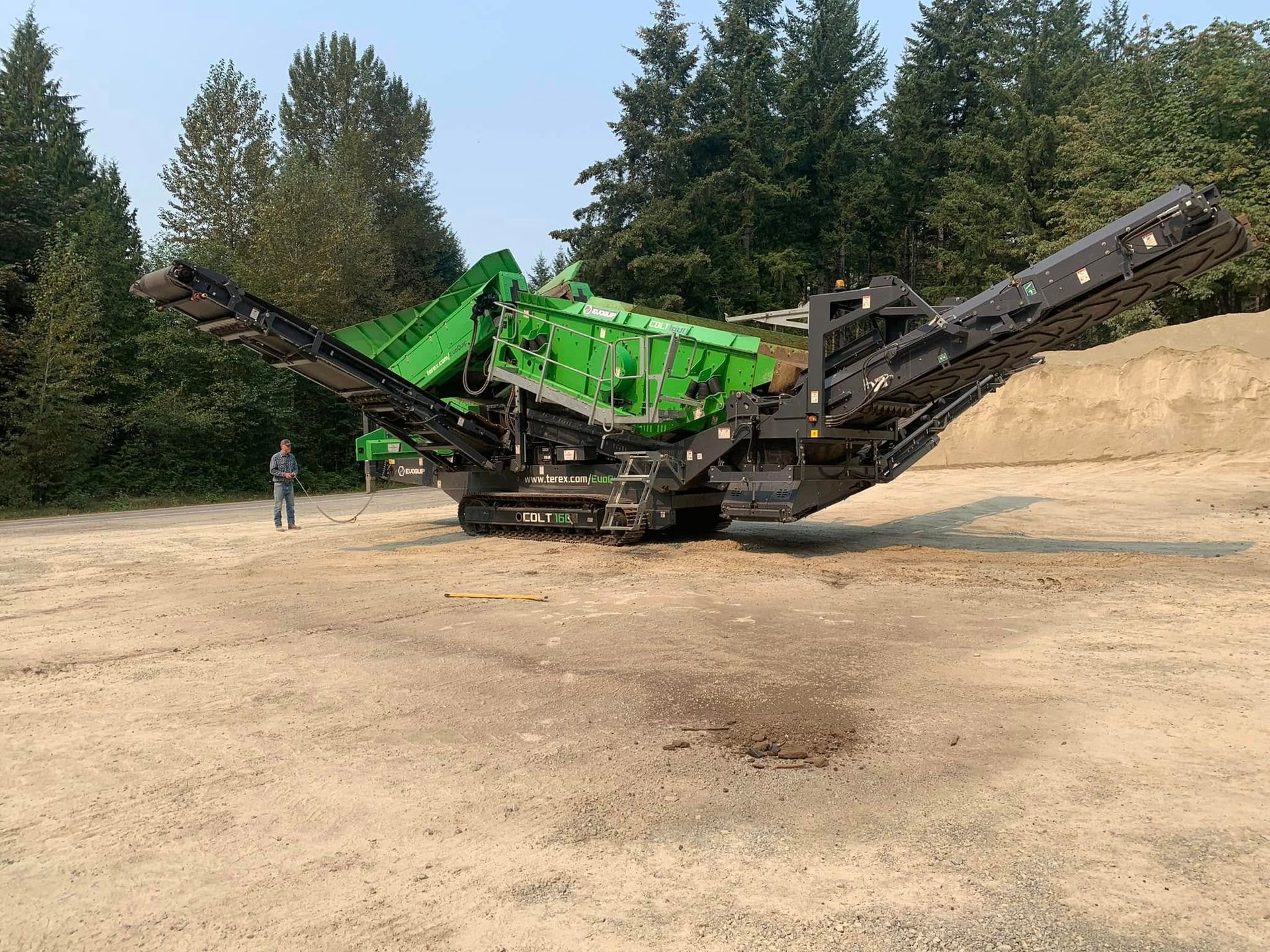 Green and black industrial rock crusher in a gravel pit with a person nearby.