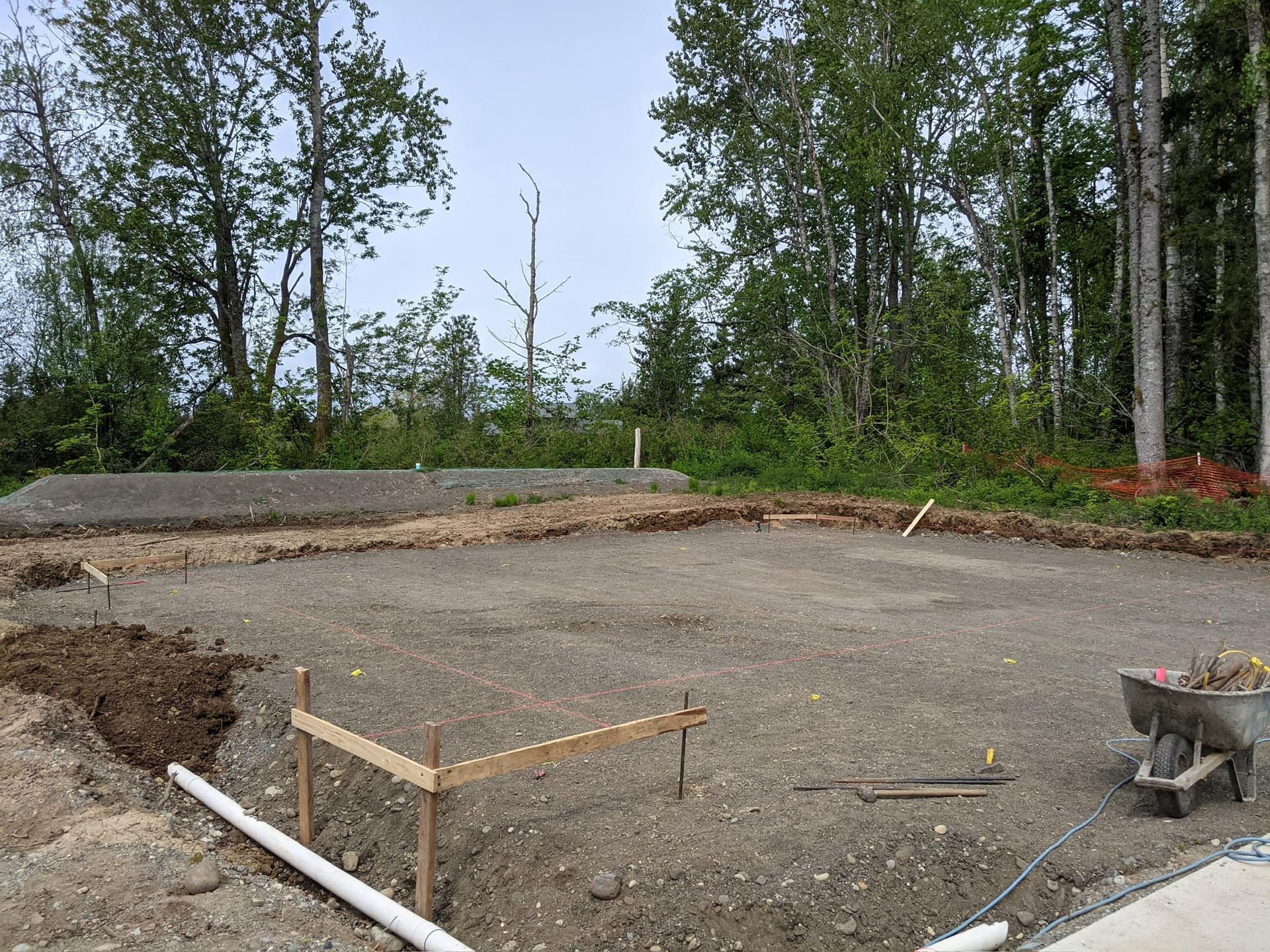 Construction site with gravel ground and wooden stakes. A small pile of dirt sits on the left. Trees in the background.