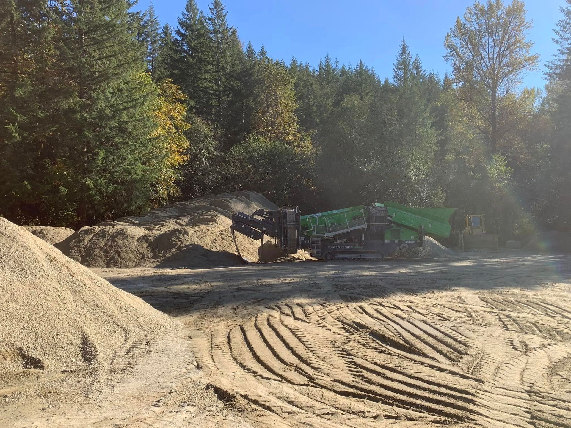 Sand piles and a dump truck in a wooded area on a sunny day.