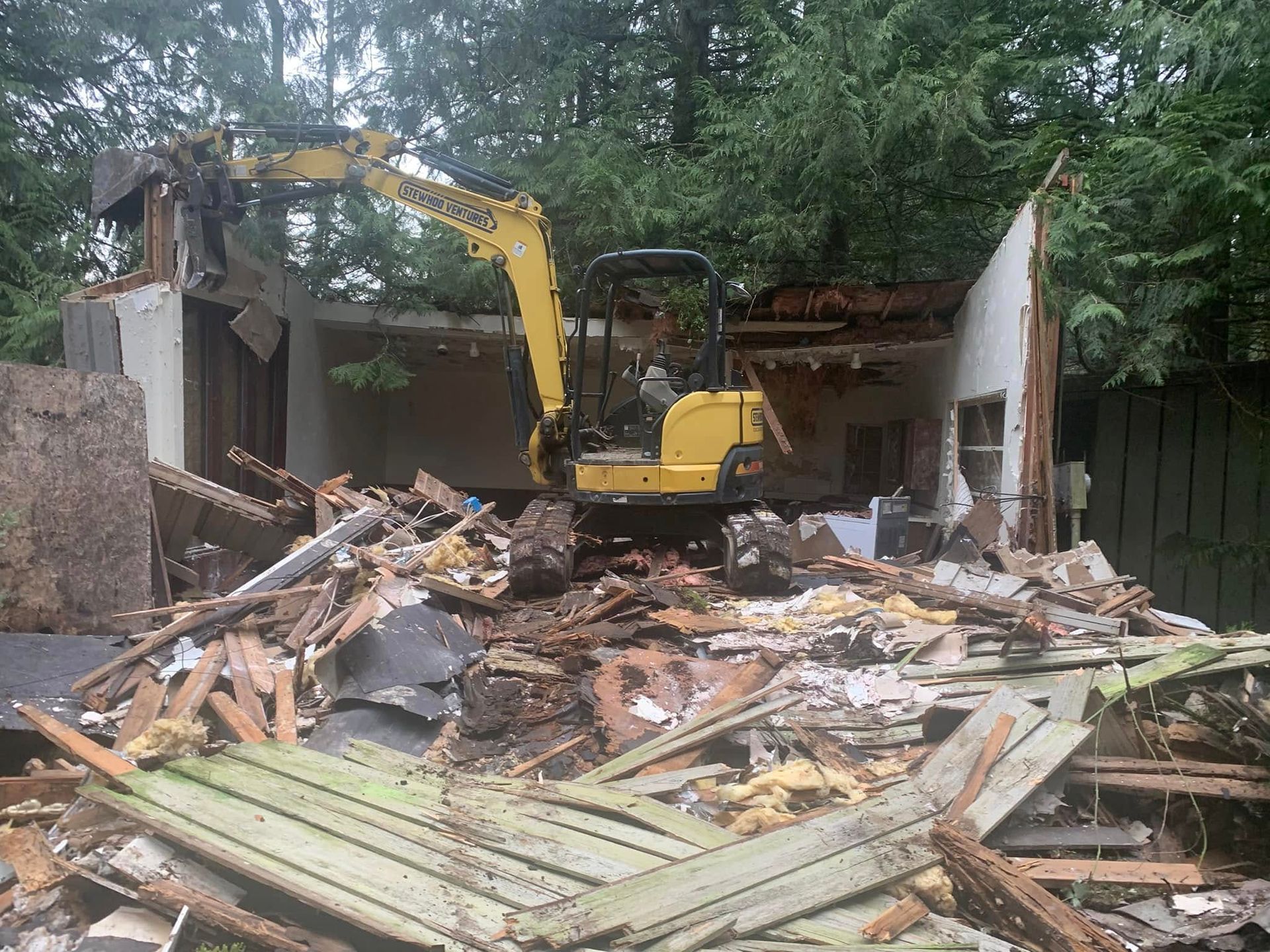 Yellow excavator demolishing a house, debris scattered, trees in background.