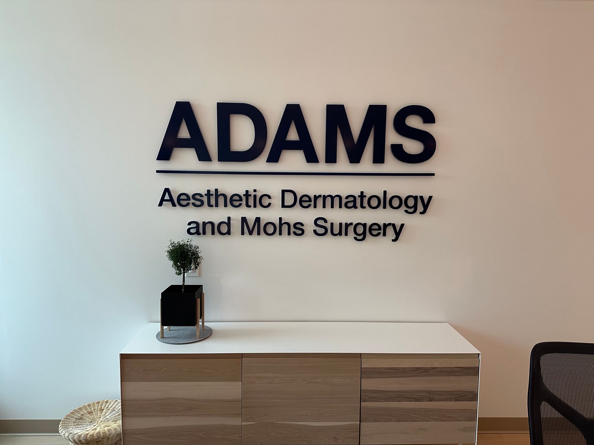 ADAMS Dermatology and Moos Surgery sign above a modern wood cabinet and plant in a clinic lobby