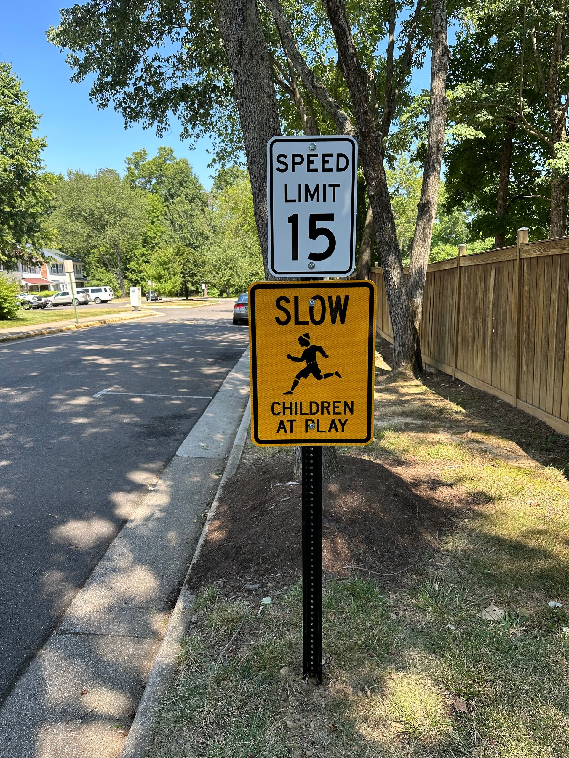 Two roadside signs: “Speed Limit 15” above yellow “SLOW Children at Play” in a leafy neighborhood.