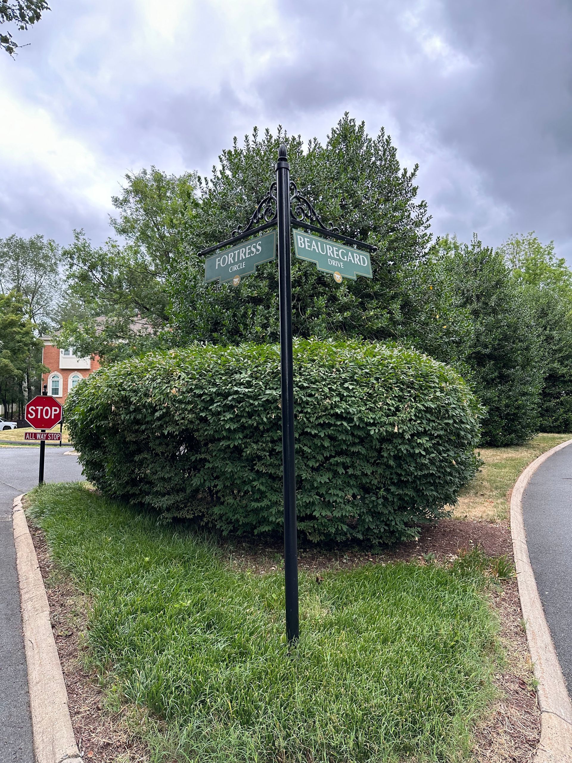 Street sign at a hedge-lined corner by a stop sign under a cloudy sky