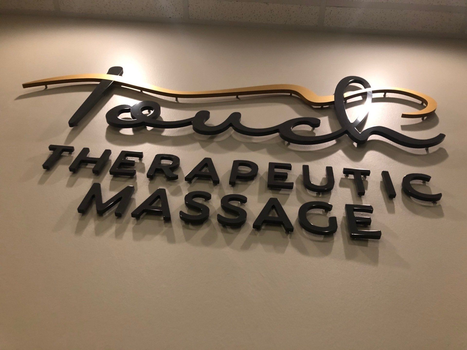Therapeutic massage sign on a beige wall, with black raised lettering and a gold cursive logo.