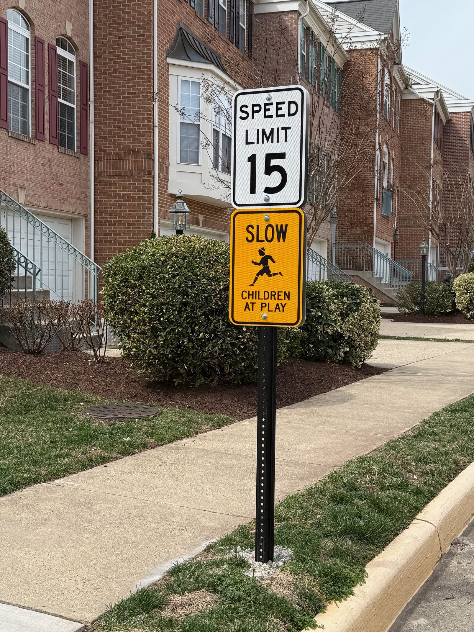 Speed limit 15 and yellow slow sign on a post beside a brick building and sidewalk.