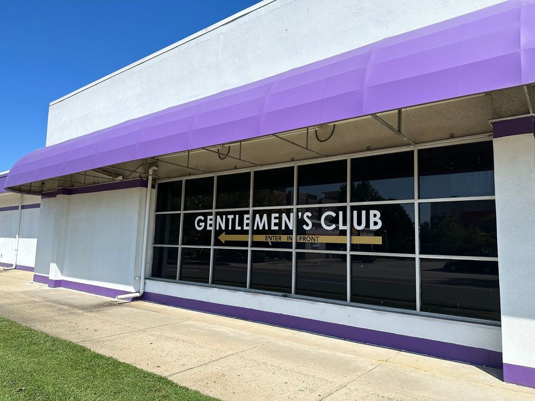 Storefront with purple awning and “Gentlemen’s Club” sign above large windows