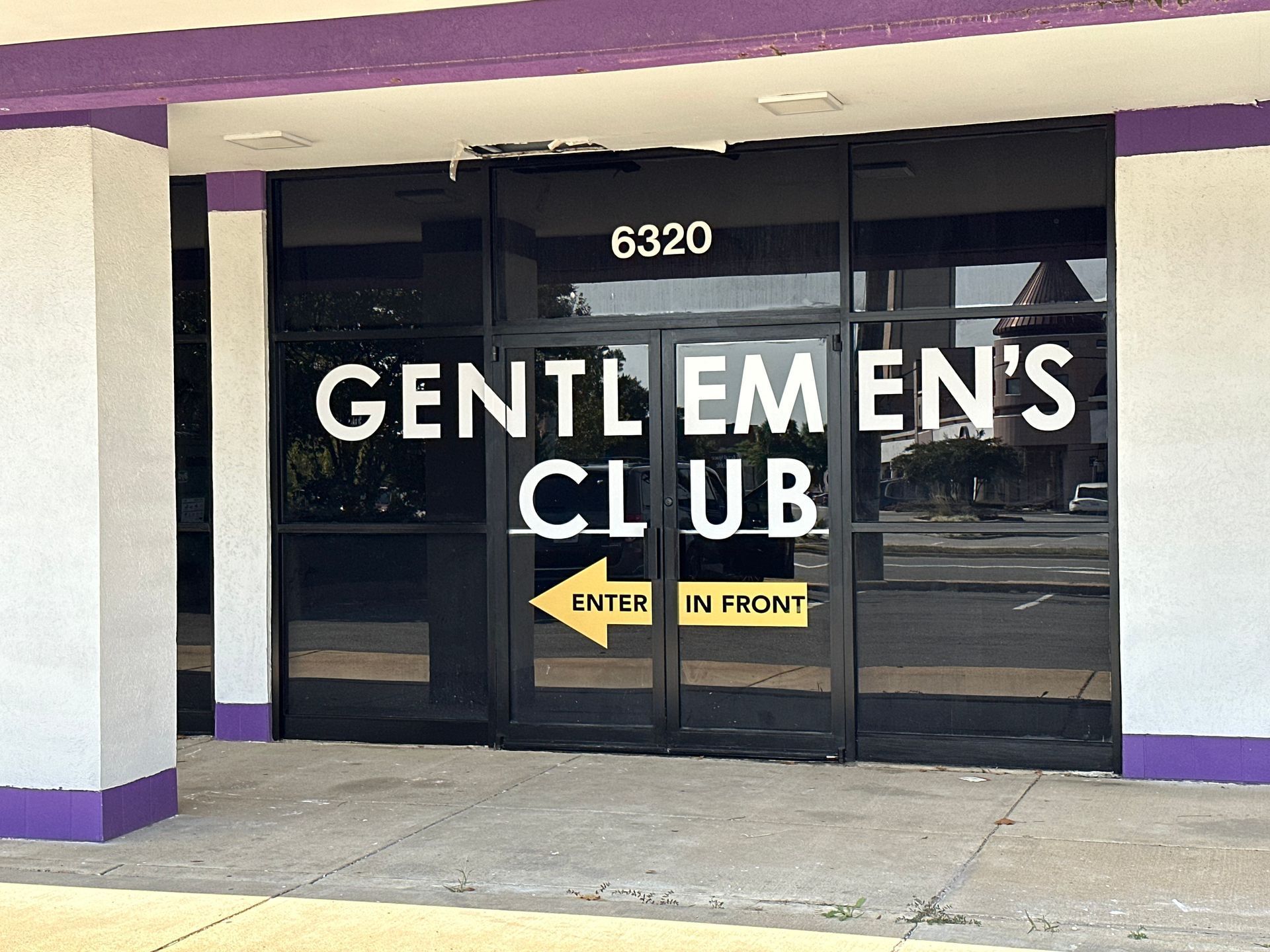 Storefront with black glass doors labeled “Gentlemen’s Club” and yellow “Must Be 18” signs.