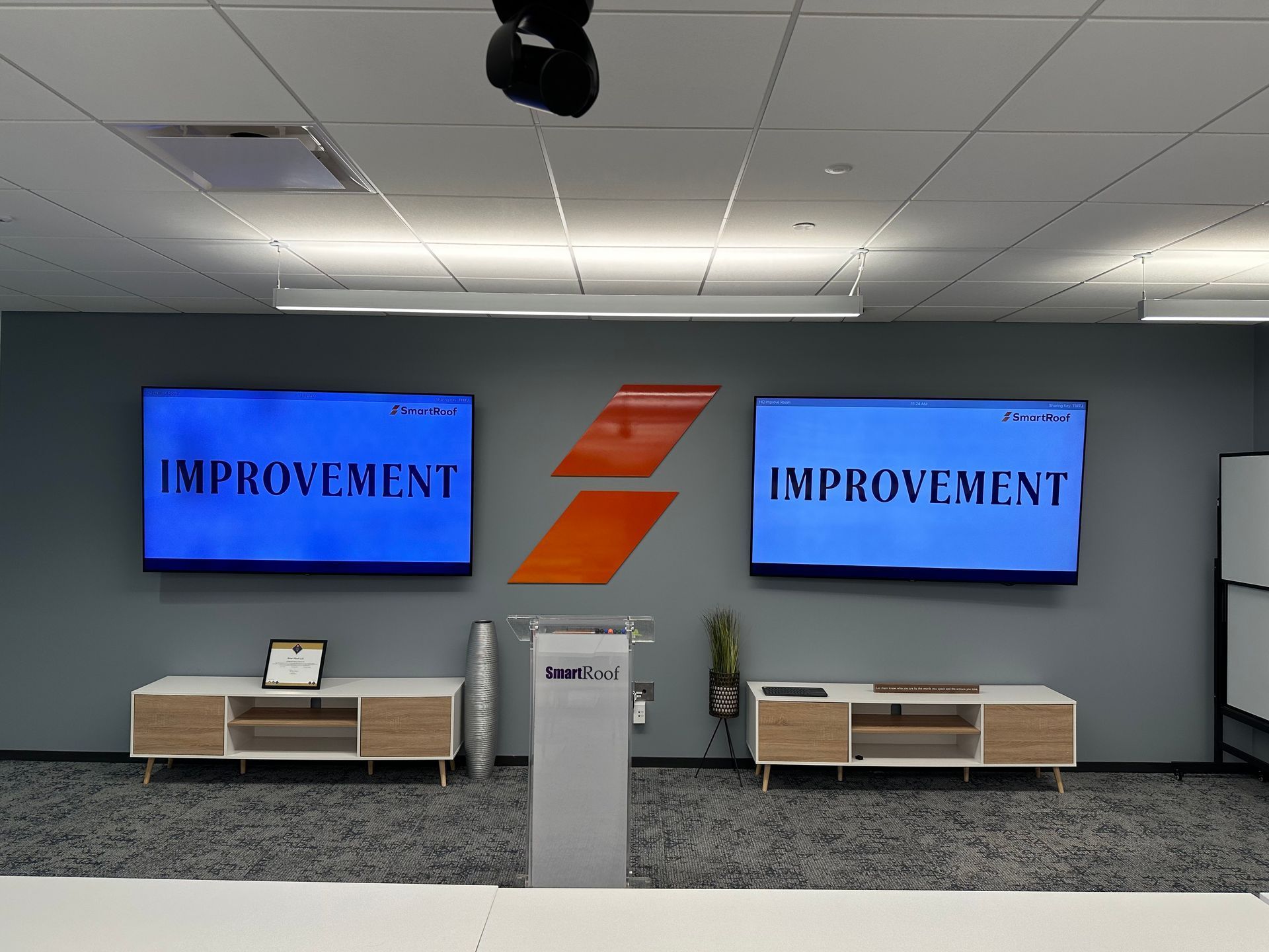 Office lobby wall with two blue screens reading “IMPROVEMENT” and an orange logo between wooden benches