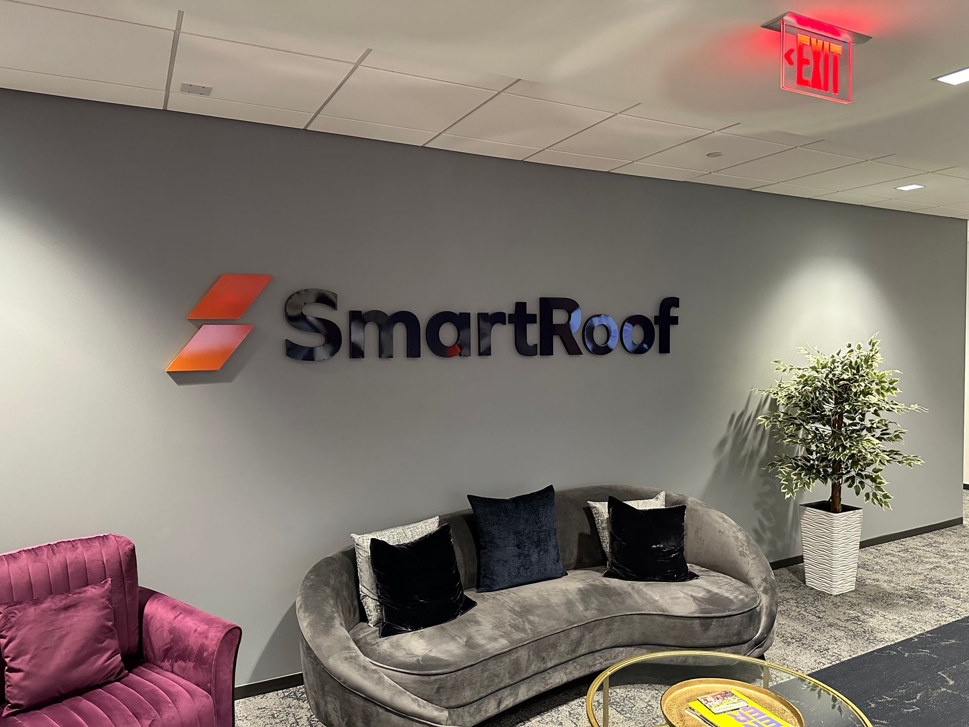 SmartRoof lobby with logo on gray wall, gray sofa, pink chair, and potted plant under ceiling lights