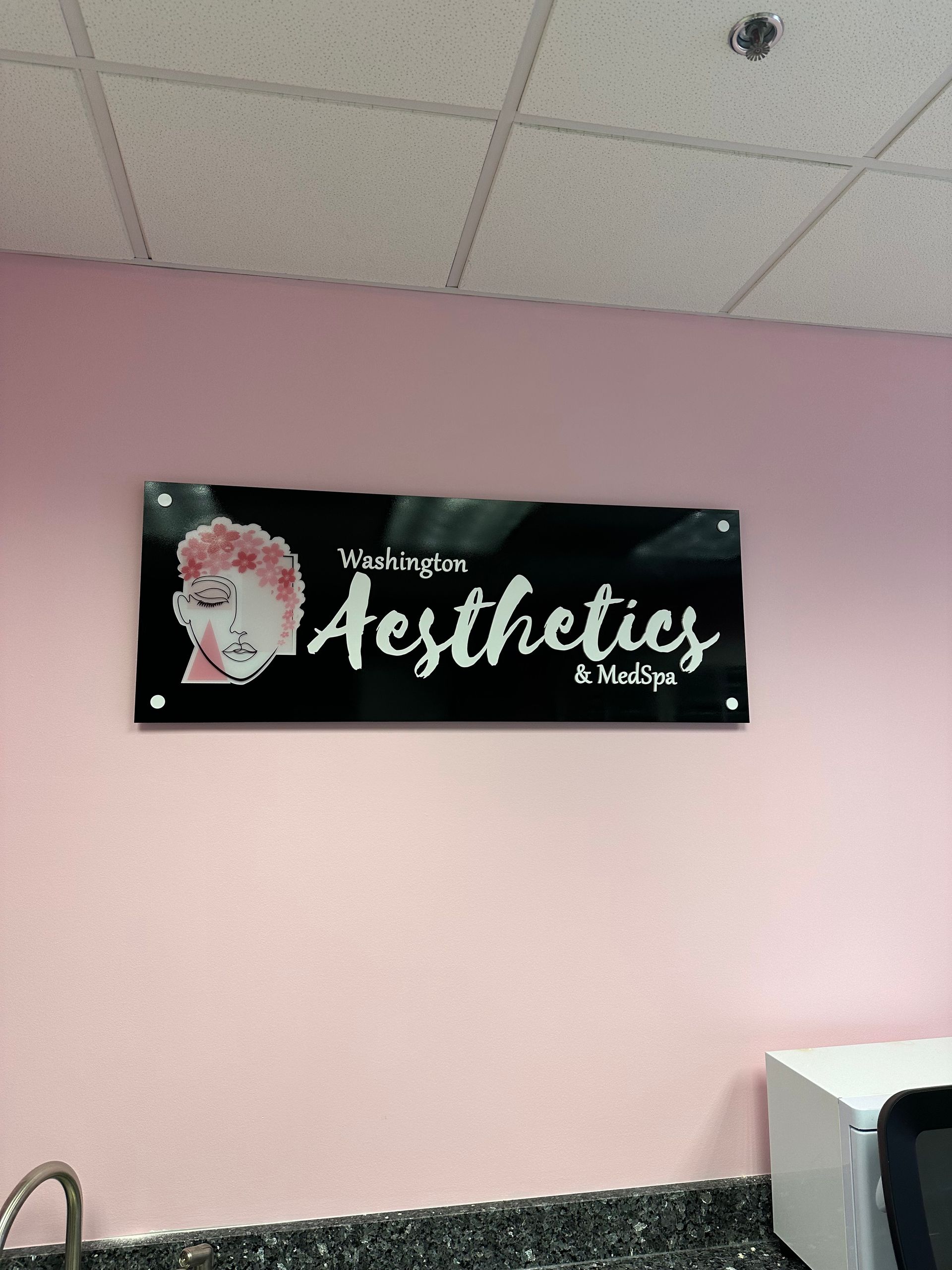 Black “Aesthetics” sign with a pink face graphic on a pale pink wall under a drop ceiling.