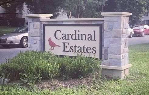 Stone entrance sign for Cardinal Estates beside greenery and parked cars