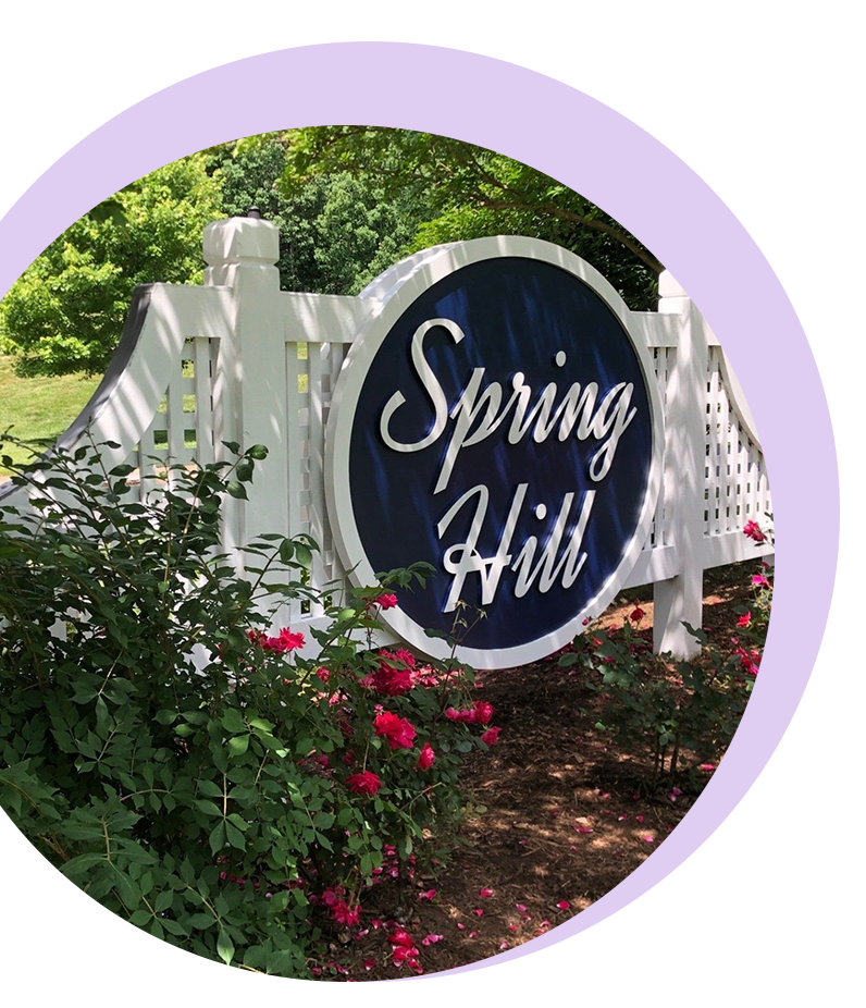 Spring Hill sign on a circular plaque amid greenery and flowers.