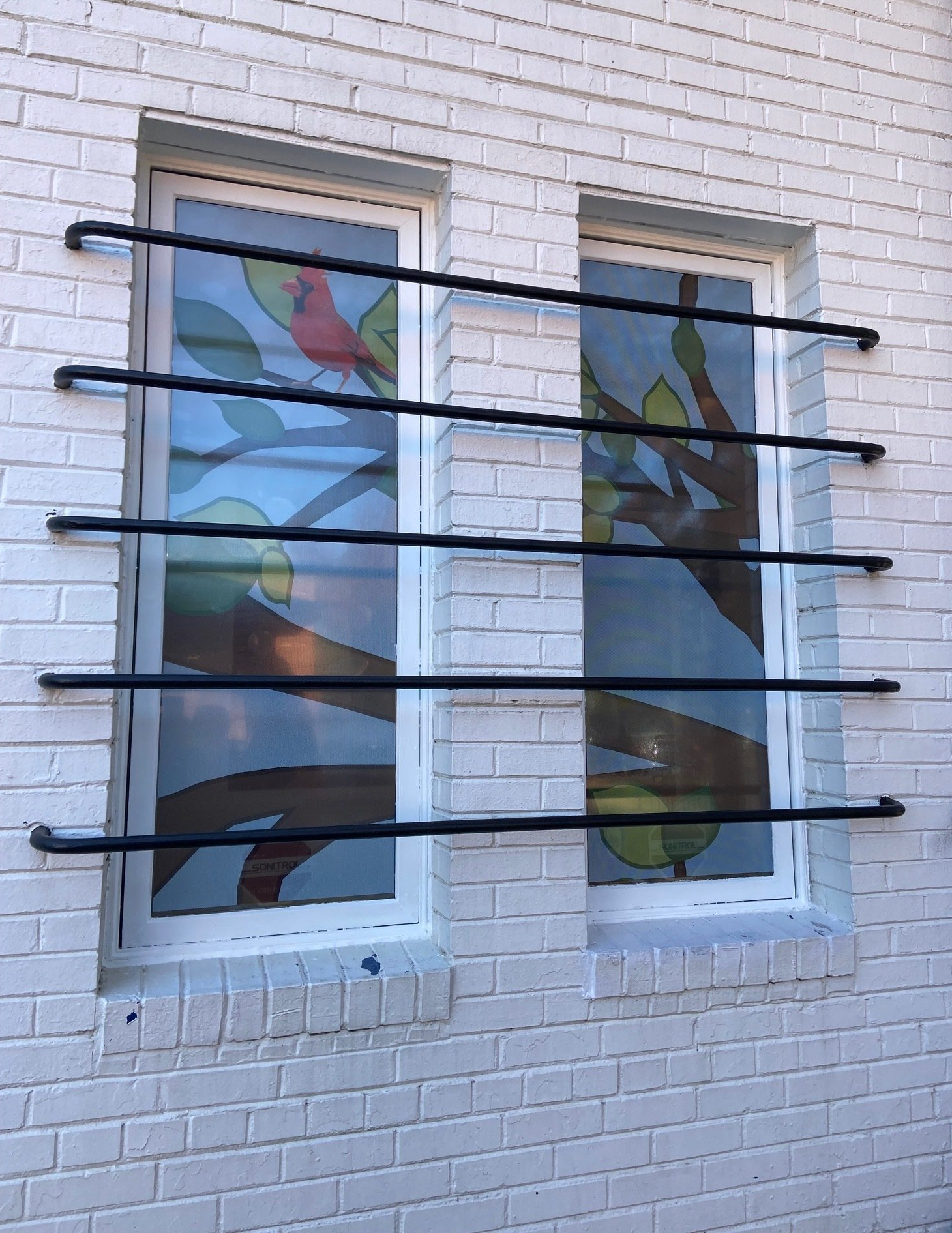 Two windows in a white brick wall, each covered with colorful stained-glass-style panels and black horizontal bars