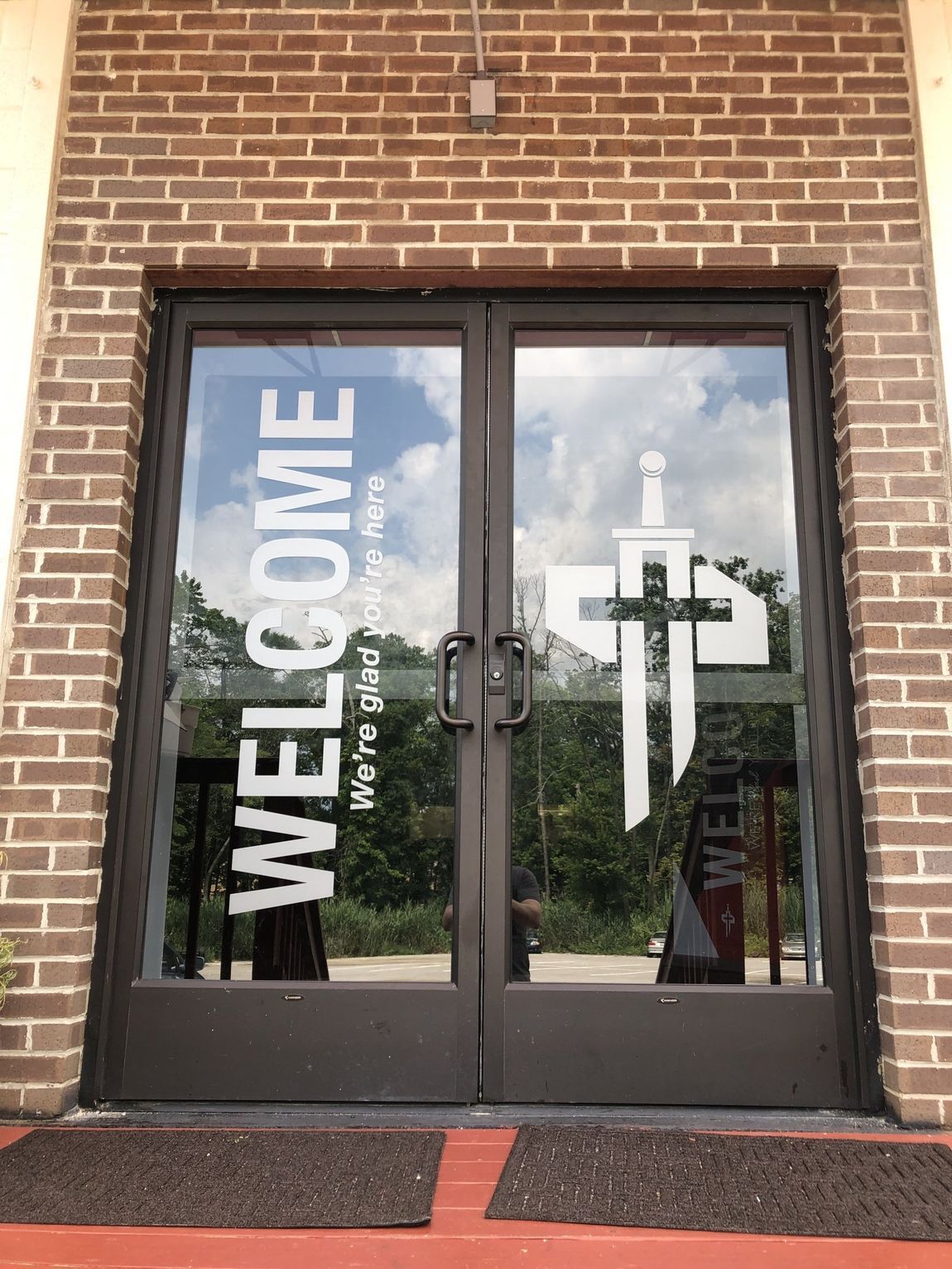Brick church entrance with glass doors reading “WELCOME” and a white cross on the right door