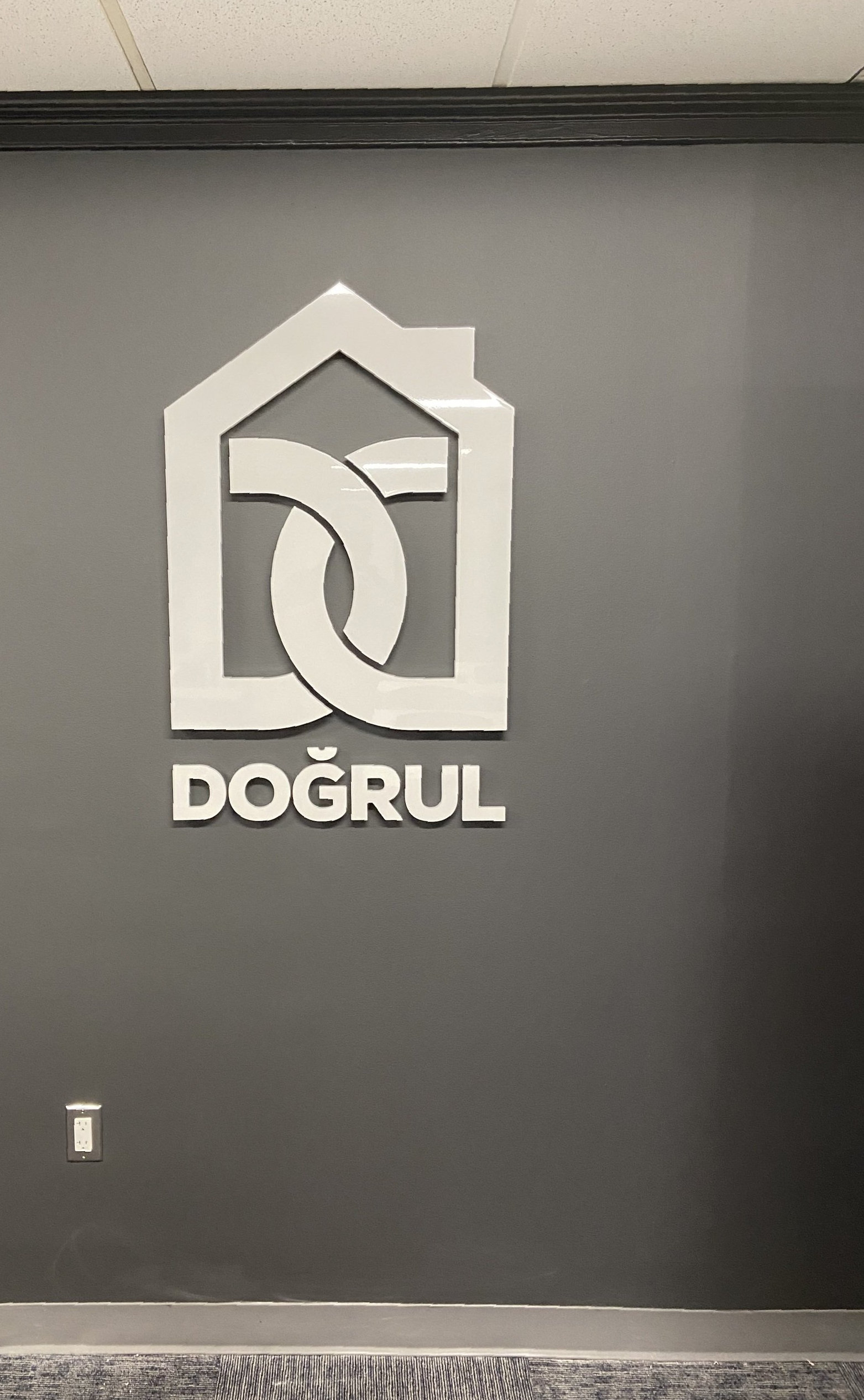 White DOĞRUL logo on a dark gray wall, shaped like a stylized house and chain link