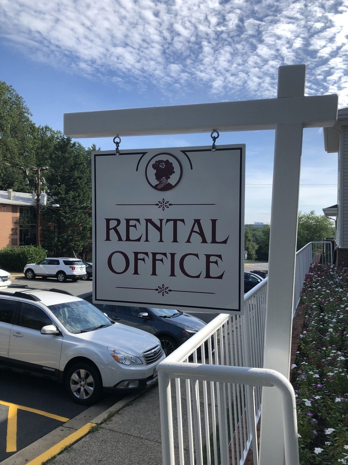 Rental office sign on a white post beside parked cars and a building, under a blue sky.