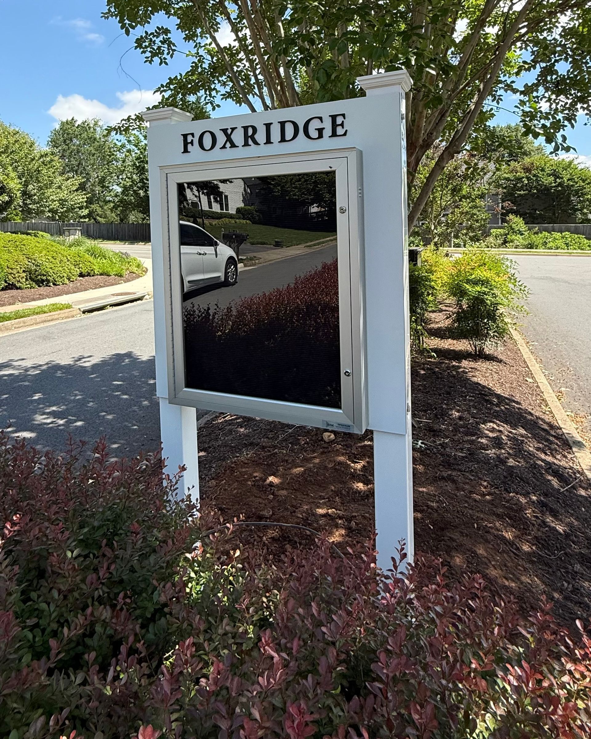 Foxridge neighborhood sign beside a road, framed by shrubs and trees on a sunny day