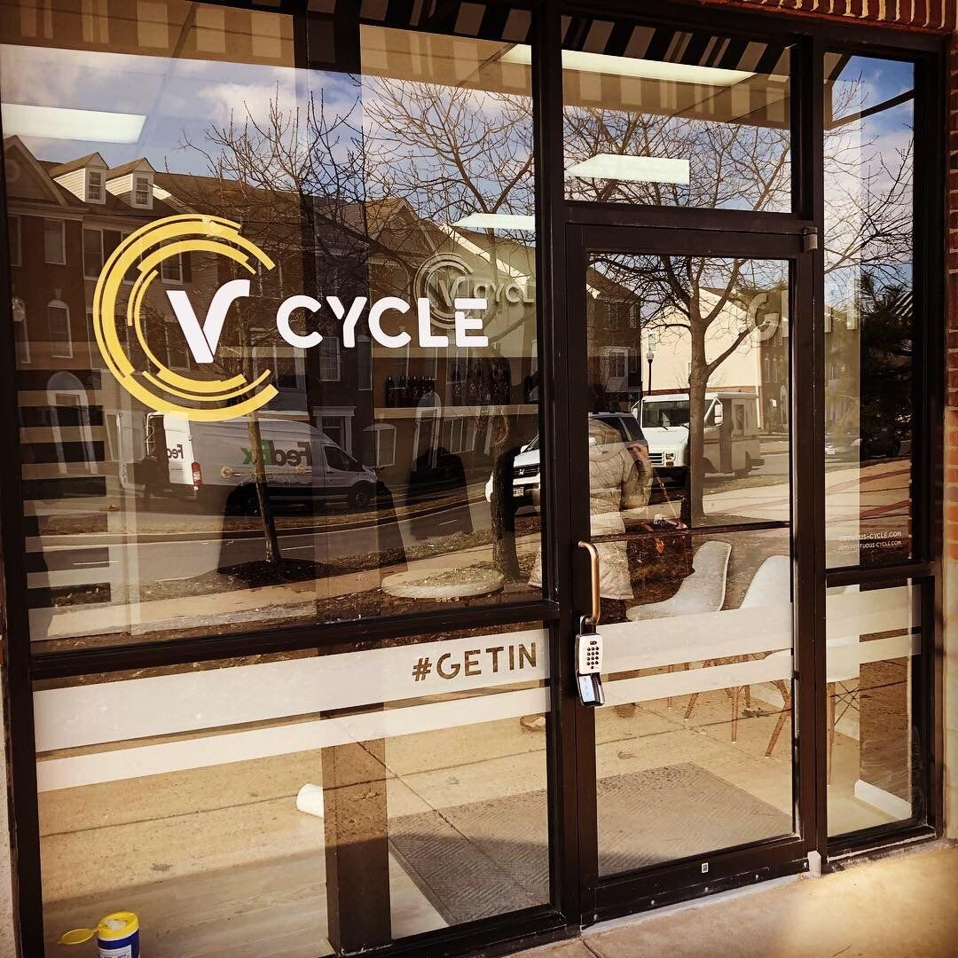 Storefront window with “V Cycle” logo and signage on a sunny street.
