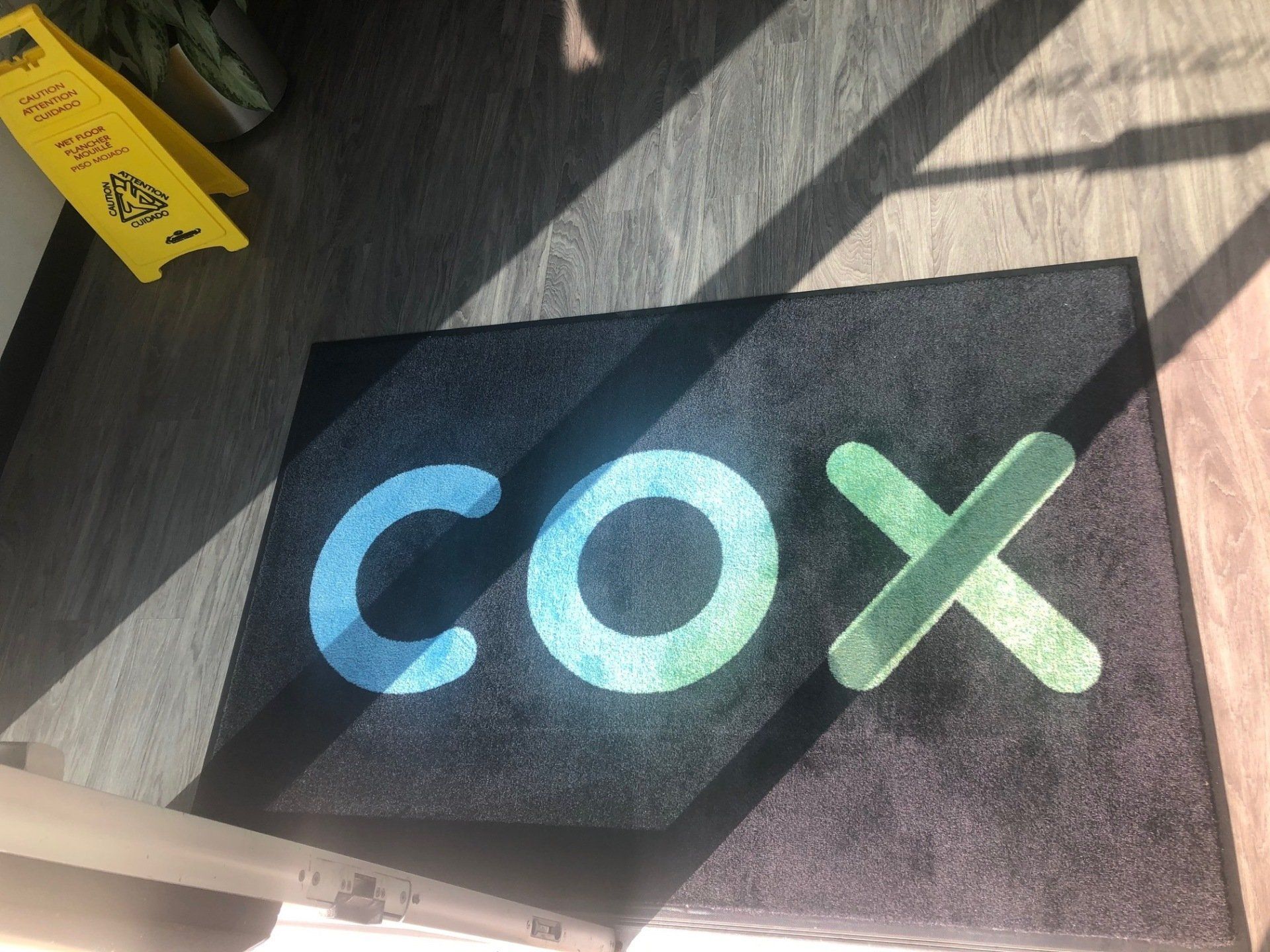 Black floor mat with large blue and green “cox” logo in sunlight on a wooden floor