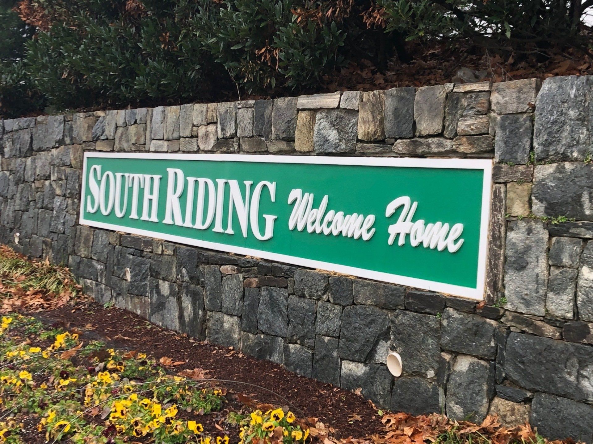 Green “South Riding Welcome Home” sign mounted on a stone wall beside flowers and shrubs
