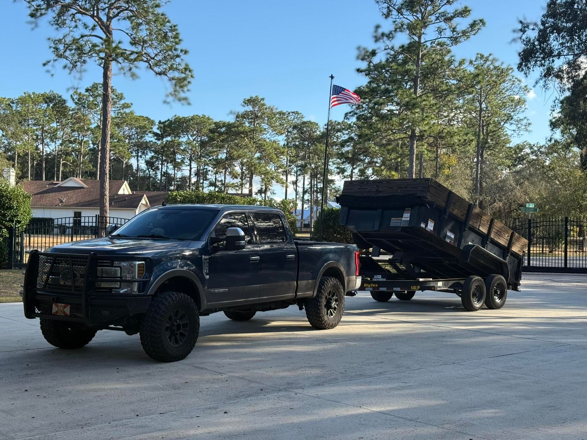A black pickup truck parked on a gravel driveway, hitched to a large, empty dump trailer against a backdrop of pine trees.