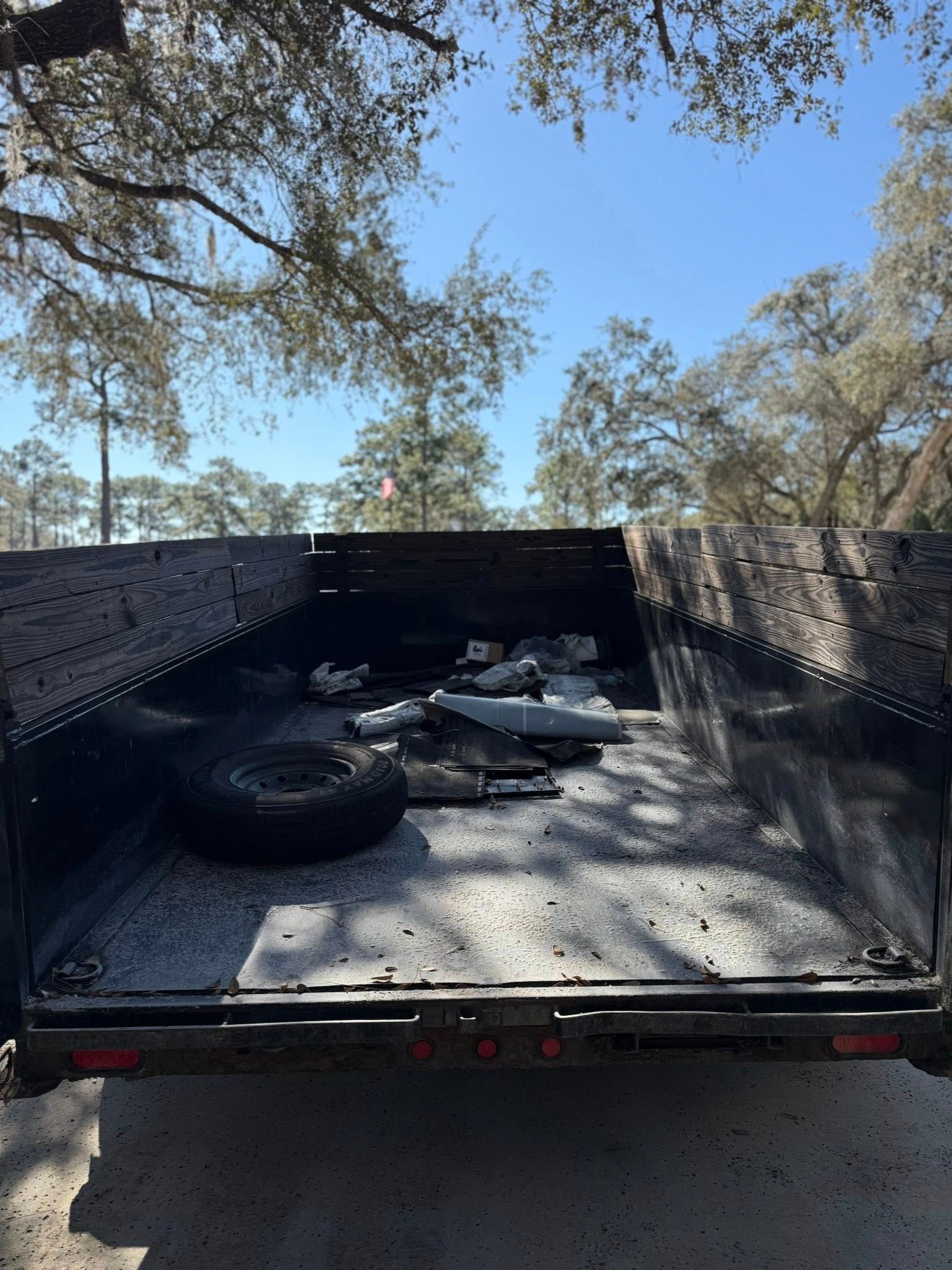 A black utility trailer bed filled with scattered debris and a spare tire, parked outdoors under trees on a sunny day.
