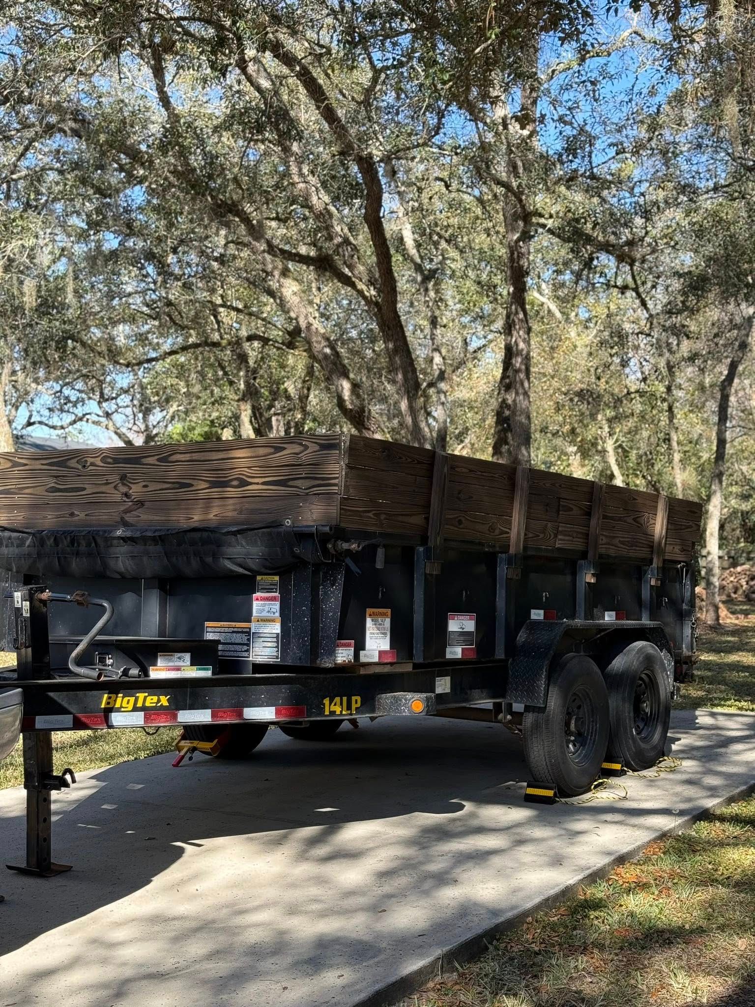 A black dump trailer parked on a concrete driveway under oak trees.