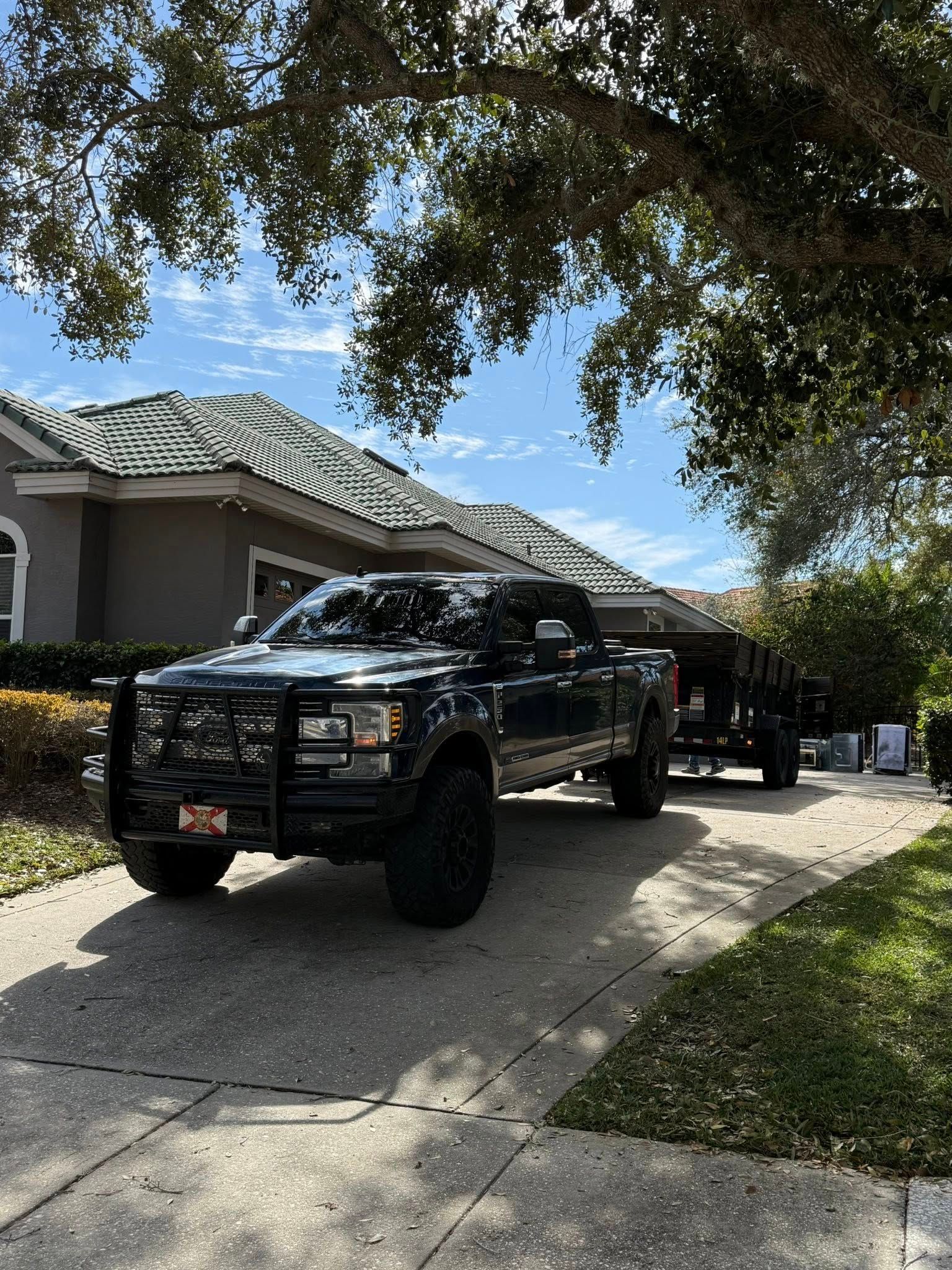 A black pickup truck with a brush guard parked on a residential driveway, hitched to a trailer in the shade of a tree.