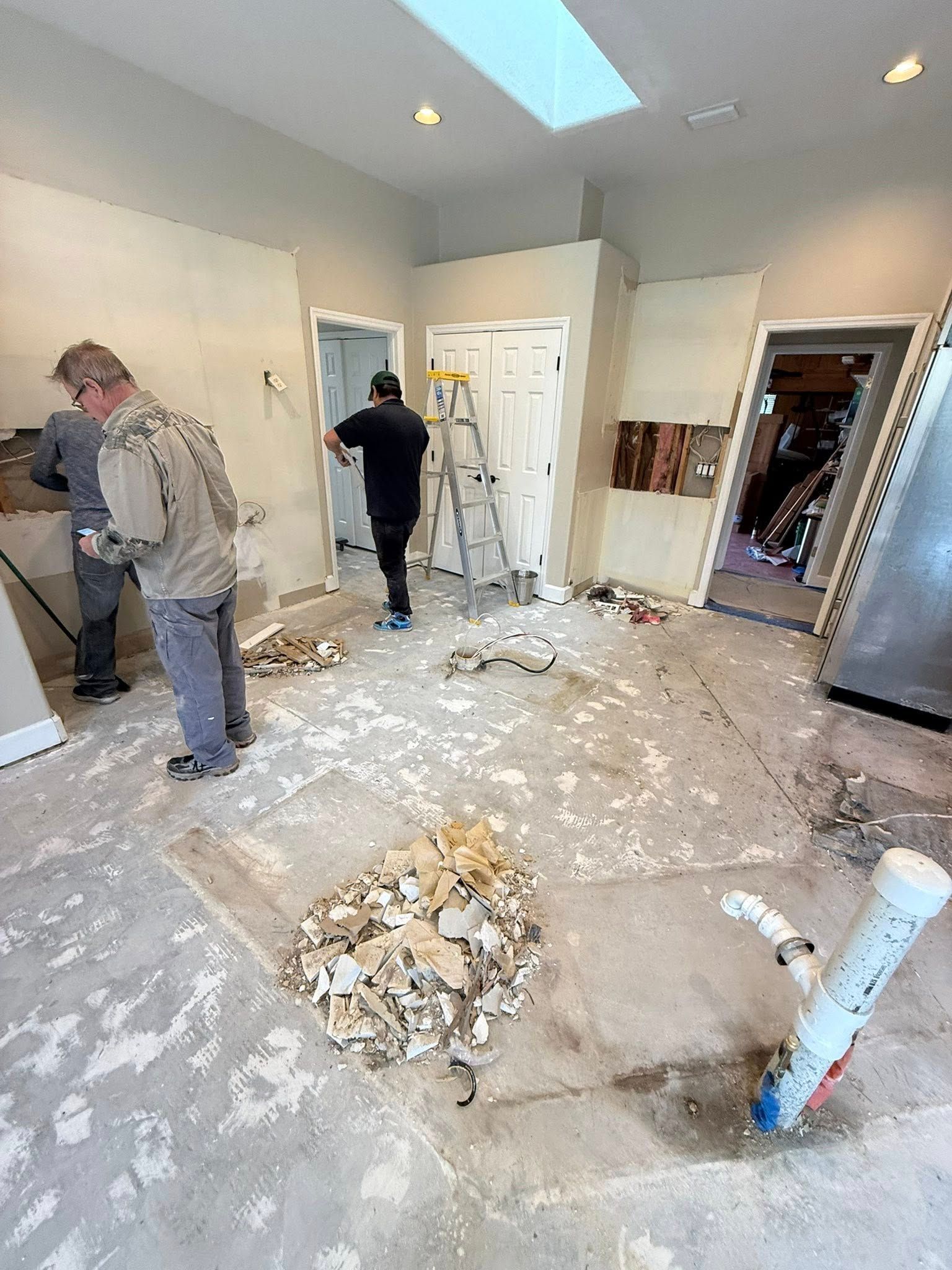 Two people renovate a room with exposed wall studs, a ladder, construction debris on the floor, and an exposed pipe.