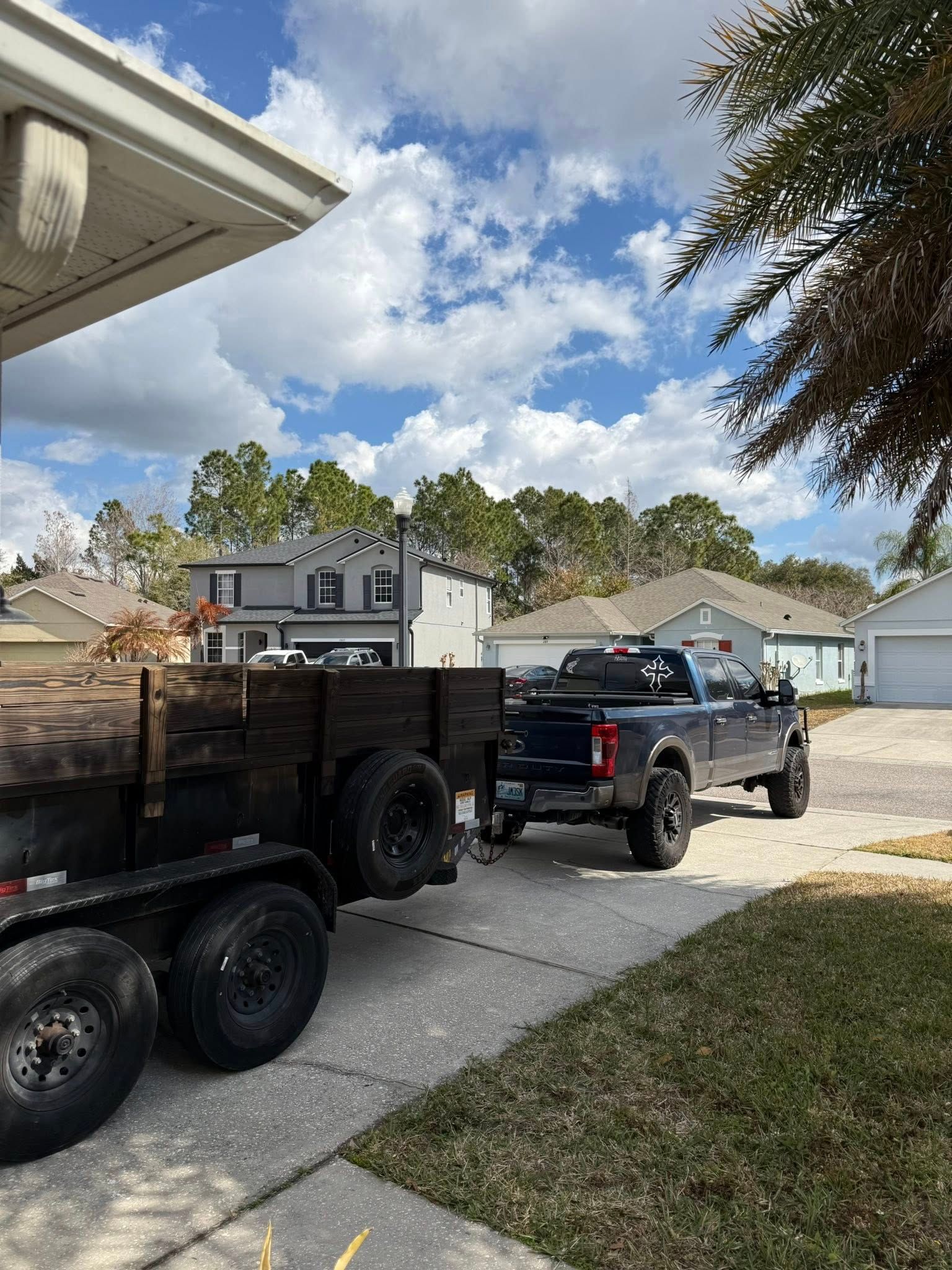 A dark pickup truck towing a flatbed trailer filled with wood logs parked in a residential driveway under a blue sky.