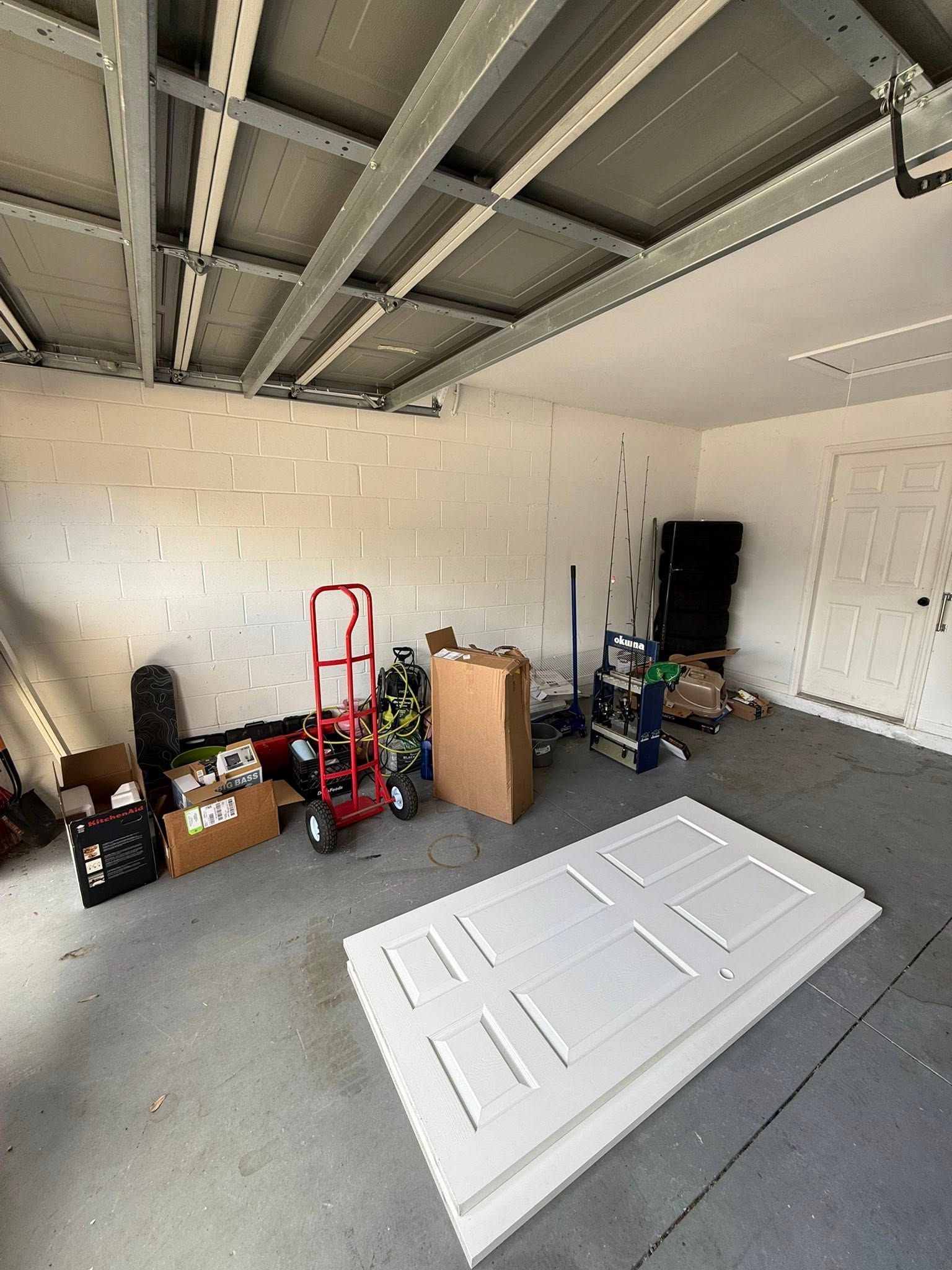 A garage with a white door lying on the gray floor, a red hand truck, a cardboard box, and miscellaneous items stored.