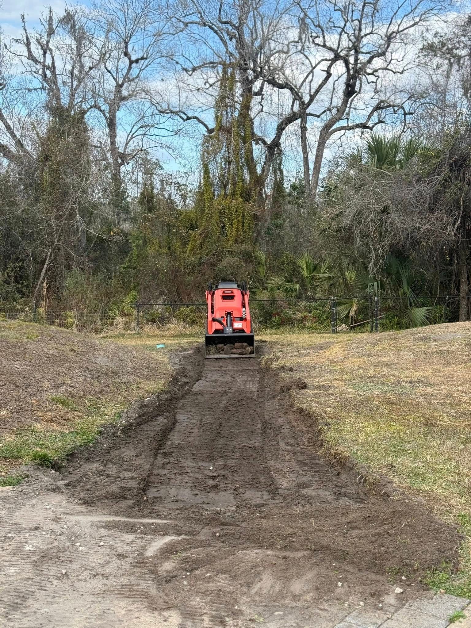 A red tiller moves away from the camera, cutting a path through dirt in a wooded, outdoor area.