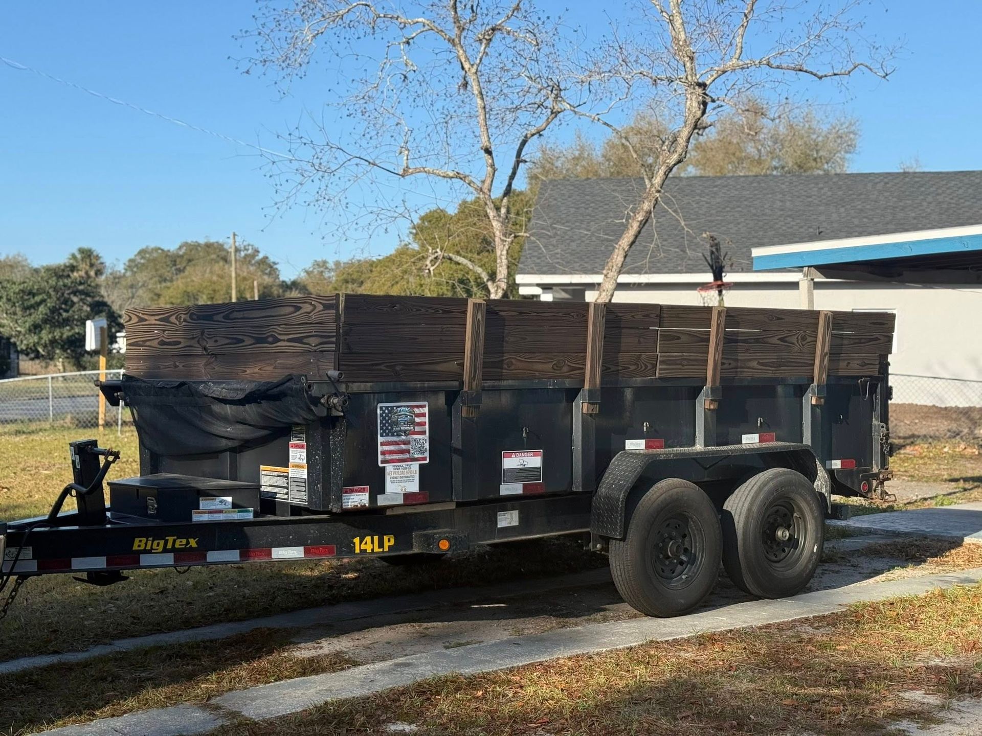 A black tandem-axle dump trailer with wooden side extensions parked on a driveway in a sunny residential area.