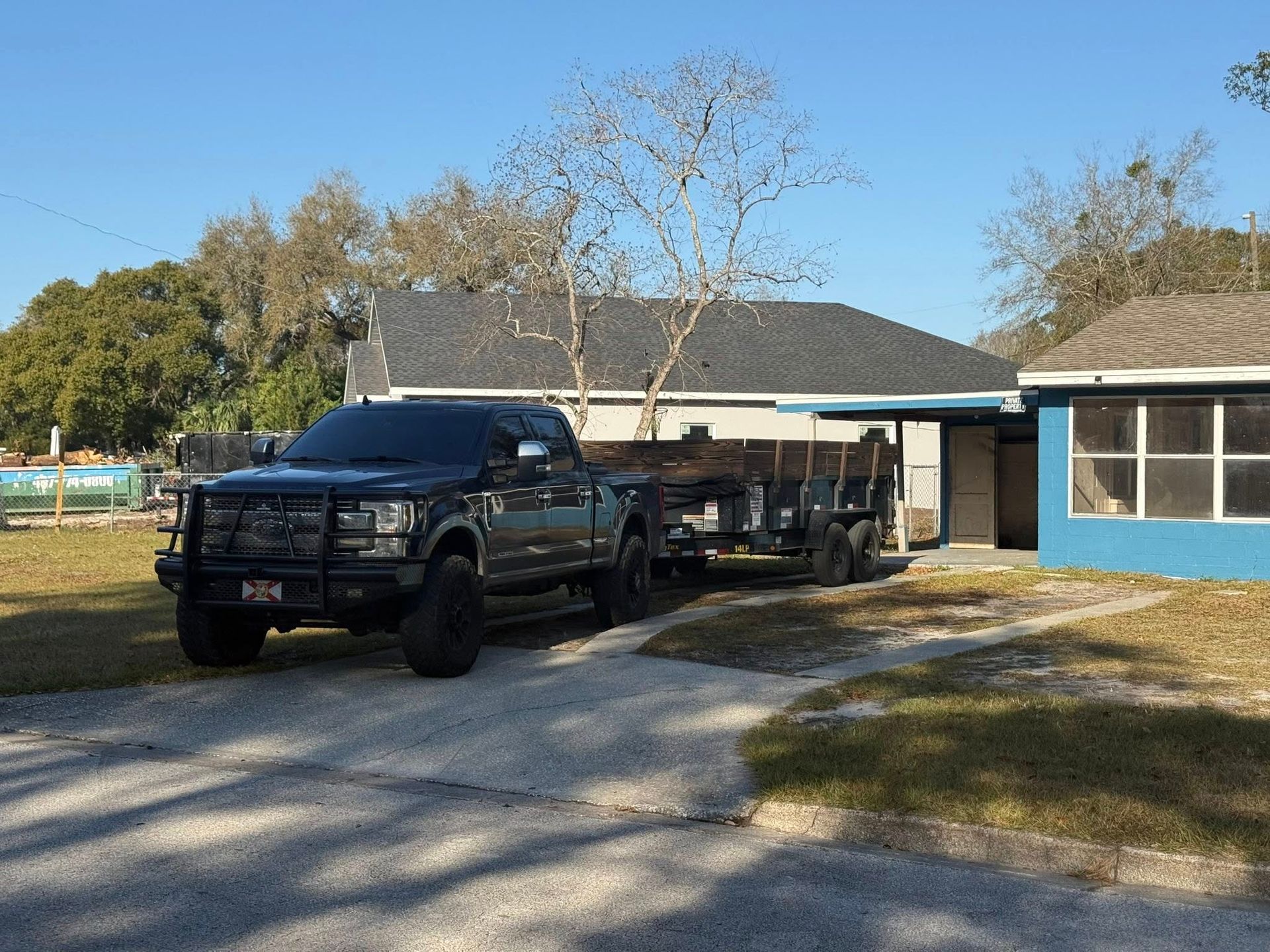 A black pickup truck with a large grille guard and an attached utility trailer parked in the gravel driveway of a house.