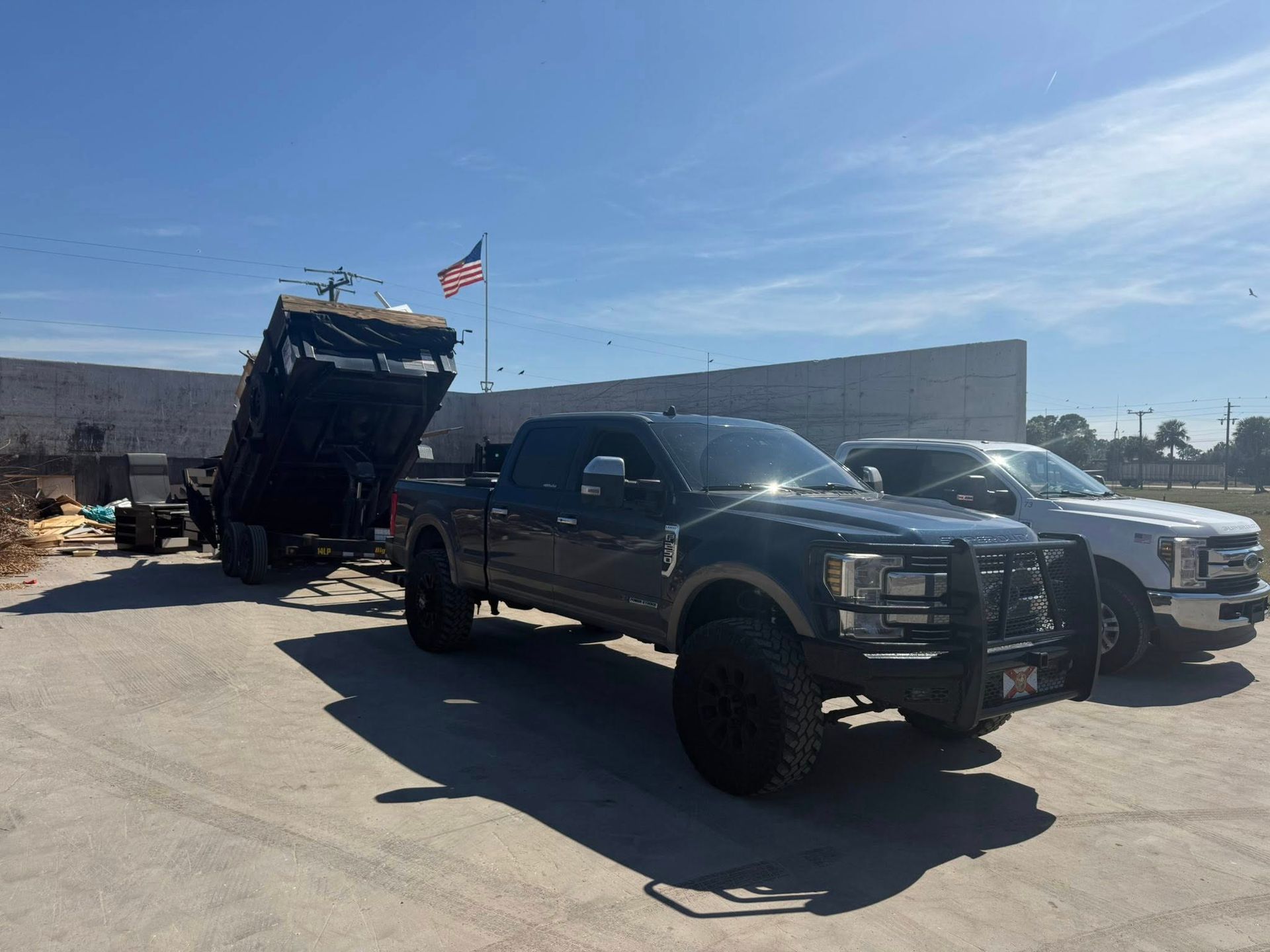 A black pickup truck towing a tilted dump trailer parked in a gravel lot under a sunny blue sky.